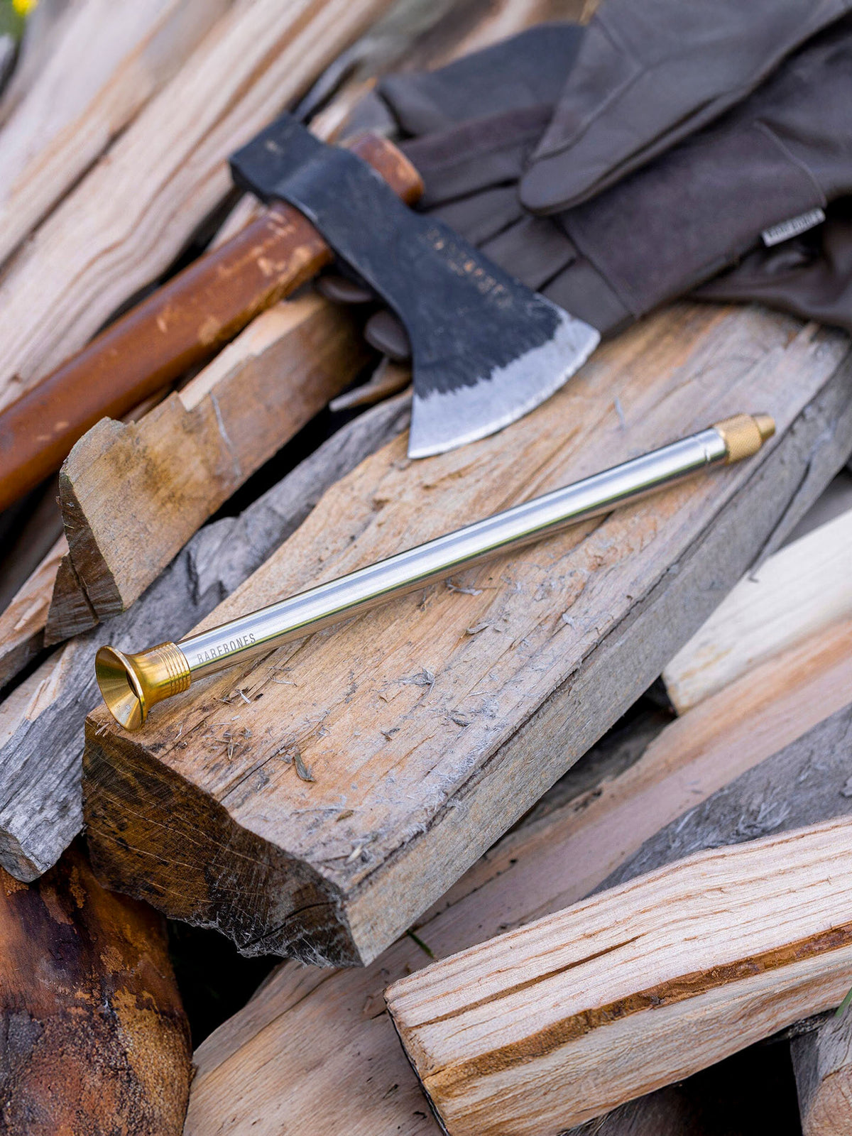 The Barebones Telescoping Fire Blower rests on a pile of split firewood, with an axe and gloves nearby.