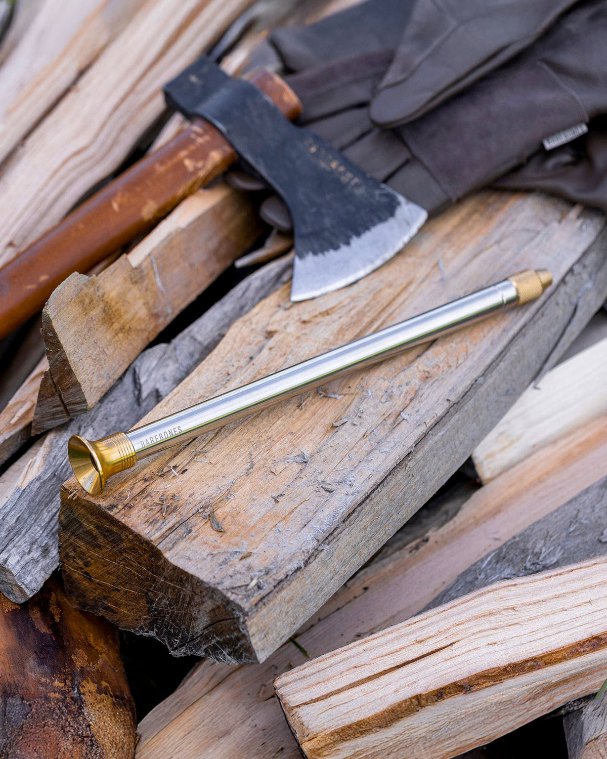 The Barebones Telescoping Fire Blower rests on a pile of split firewood, with an axe and gloves nearby.