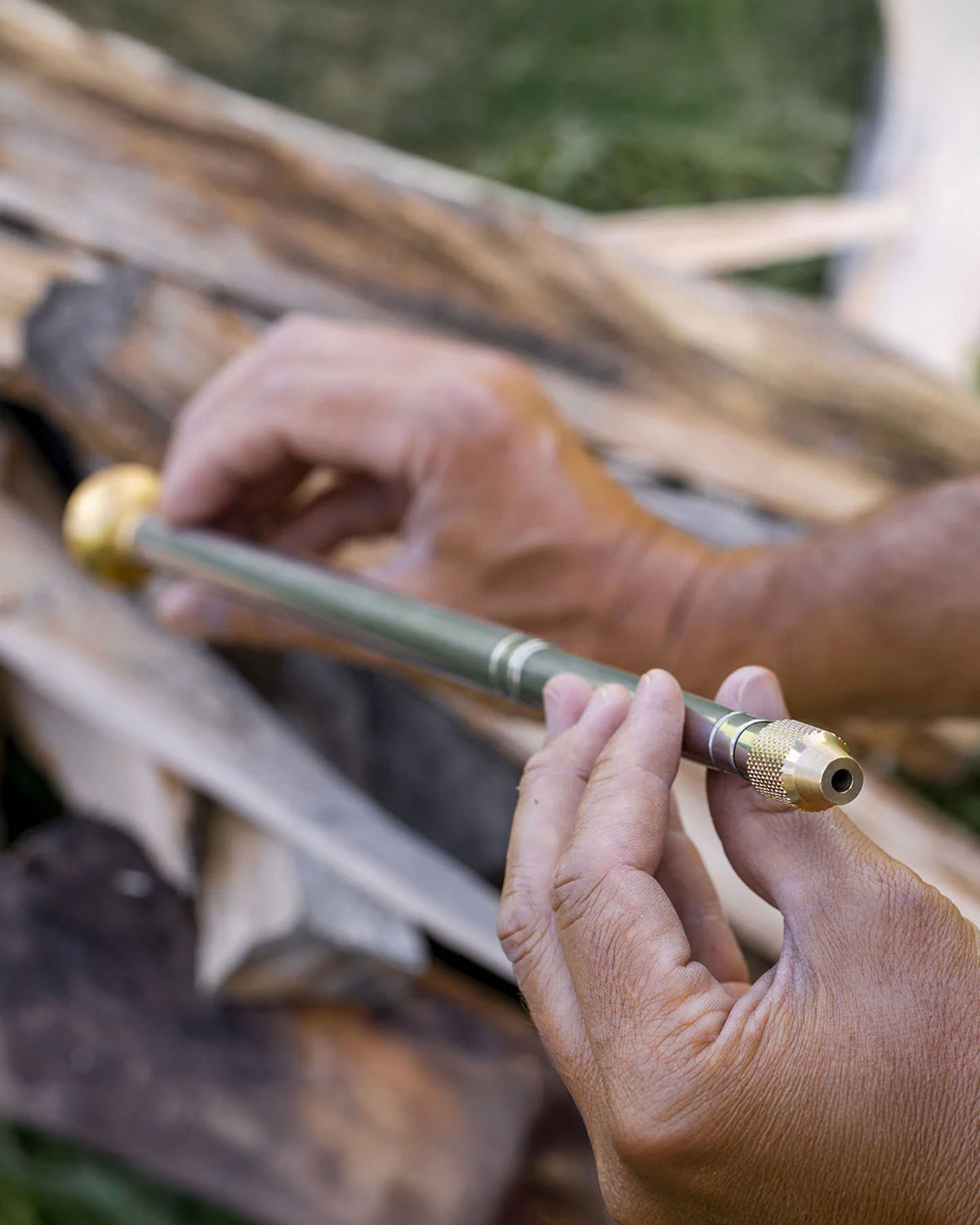 A person holds the Barebones Telescoping Fire Blower, featuring brass ends and a green body, with split firewood visible in the background.