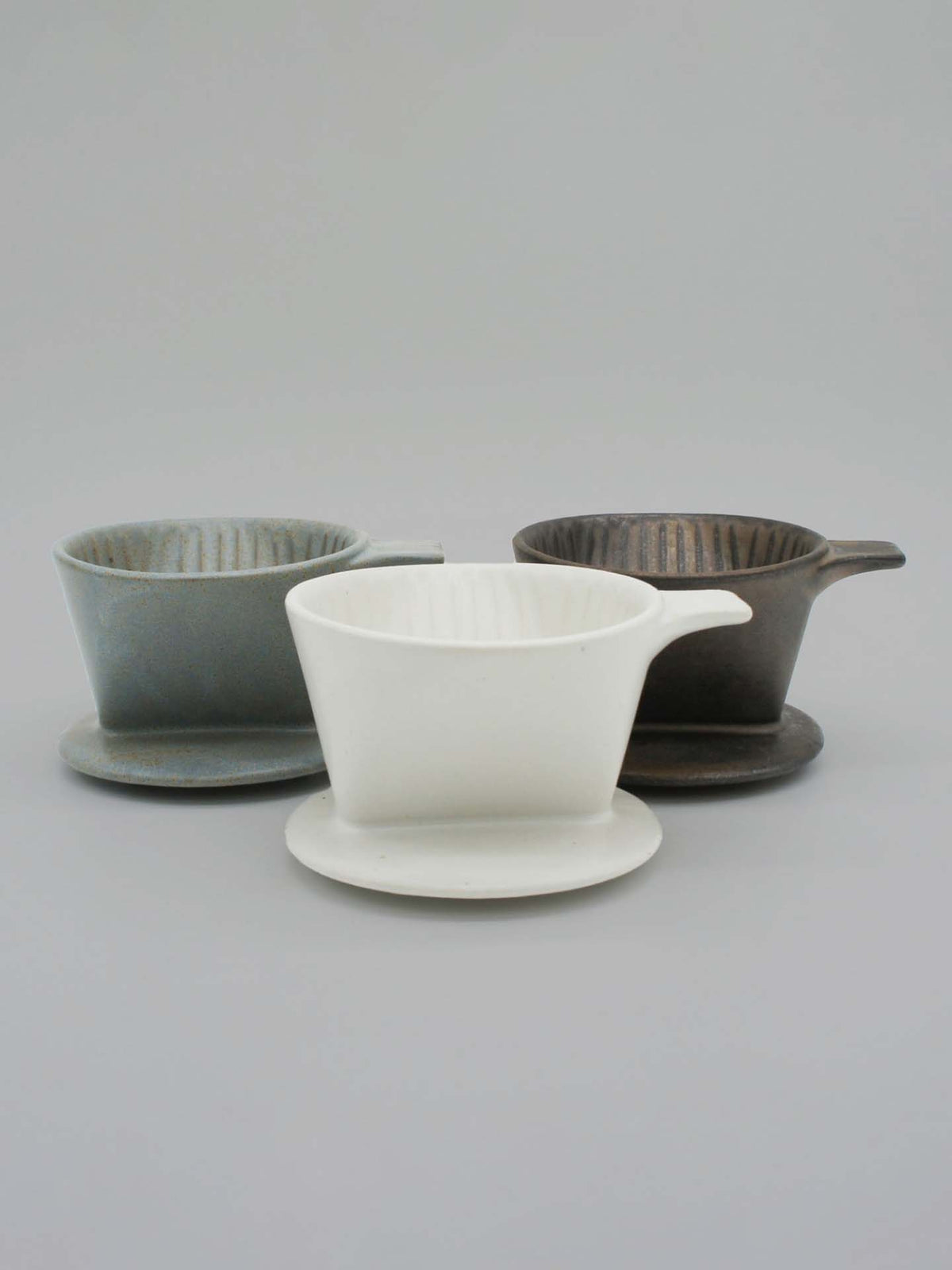 Three Mino-yaki Coffee Drippers (2 Cup) in gray, white, and dark brown from CHIPS Inc. are elegantly displayed on a light gray surface, highlighting modern craftsmanship blended with the refined elegance of traditional pottery.