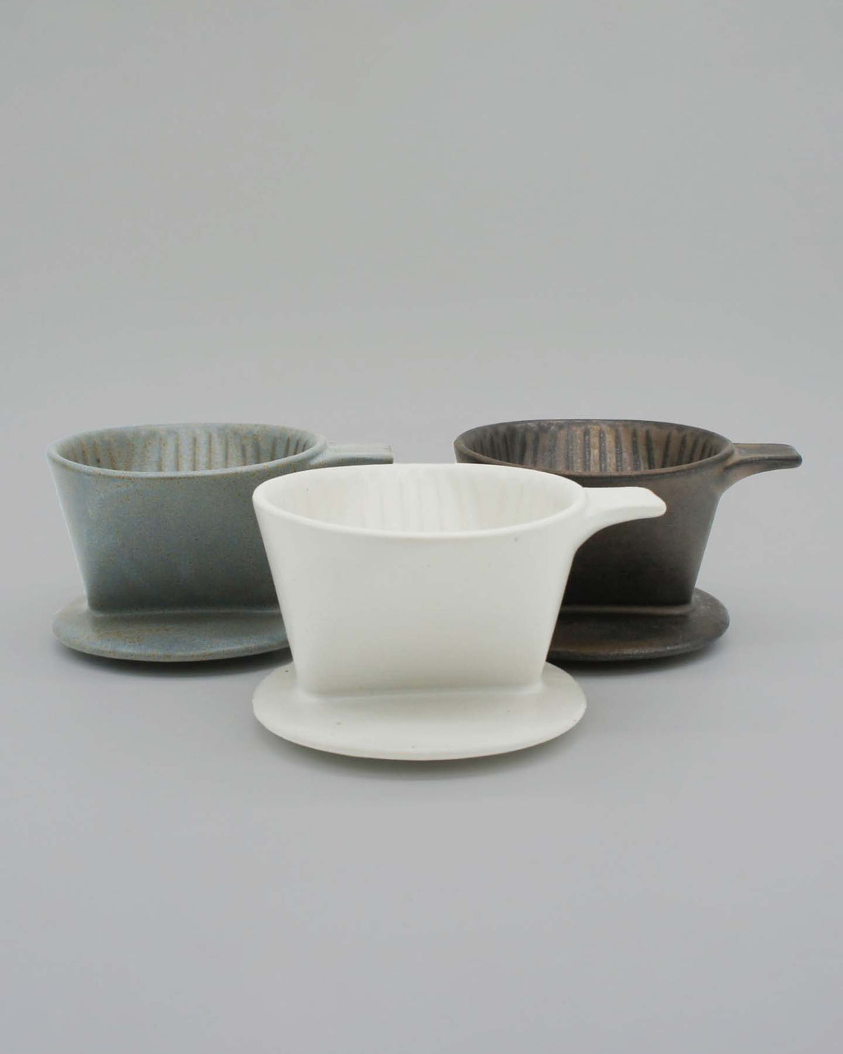 Three Mino-yaki Coffee Drippers (2 Cup) in gray, white, and dark brown from CHIPS Inc. are elegantly displayed on a light gray surface, highlighting modern craftsmanship blended with the refined elegance of traditional pottery.