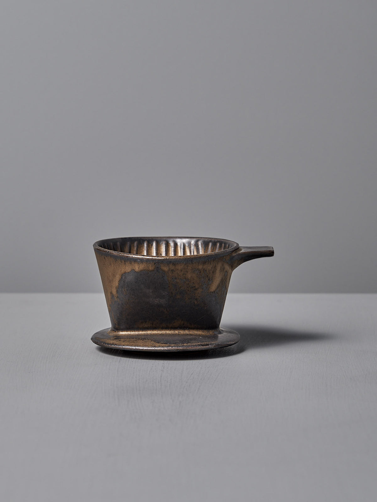 A Mino-yaki Coffee Dripper (2 Cup) – Brass by CHIPS Inc., with a small handle, rests on a matching saucer against a plain gray background.