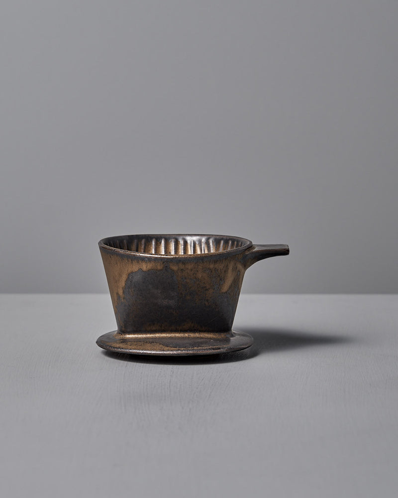 A Mino-yaki Coffee Dripper (2 Cup) – Brass by CHIPS Inc., with a small handle, rests on a matching saucer against a plain gray background.