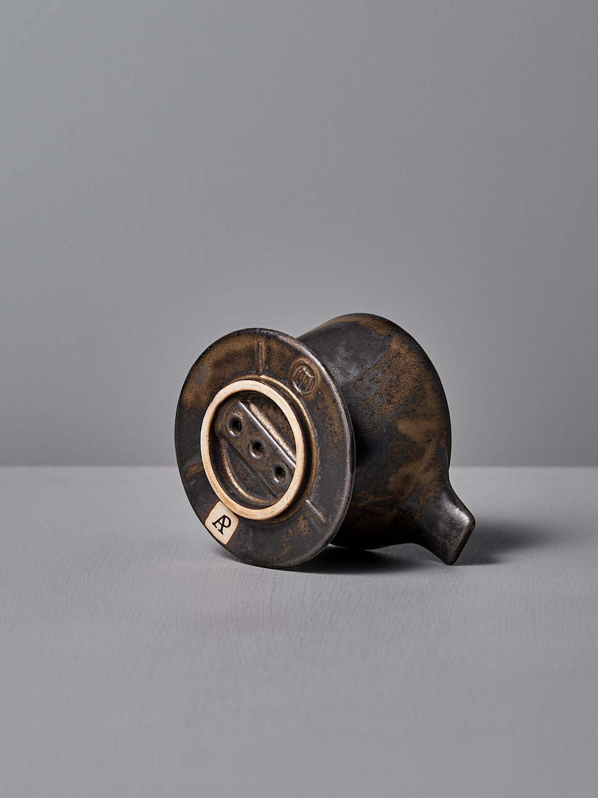 A small, vintage Mino-yaki Coffee Dripper (2 Cup) – Brass by CHIPS Inc. lies on its side revealing its round base and drainage holes against a plain gray background.