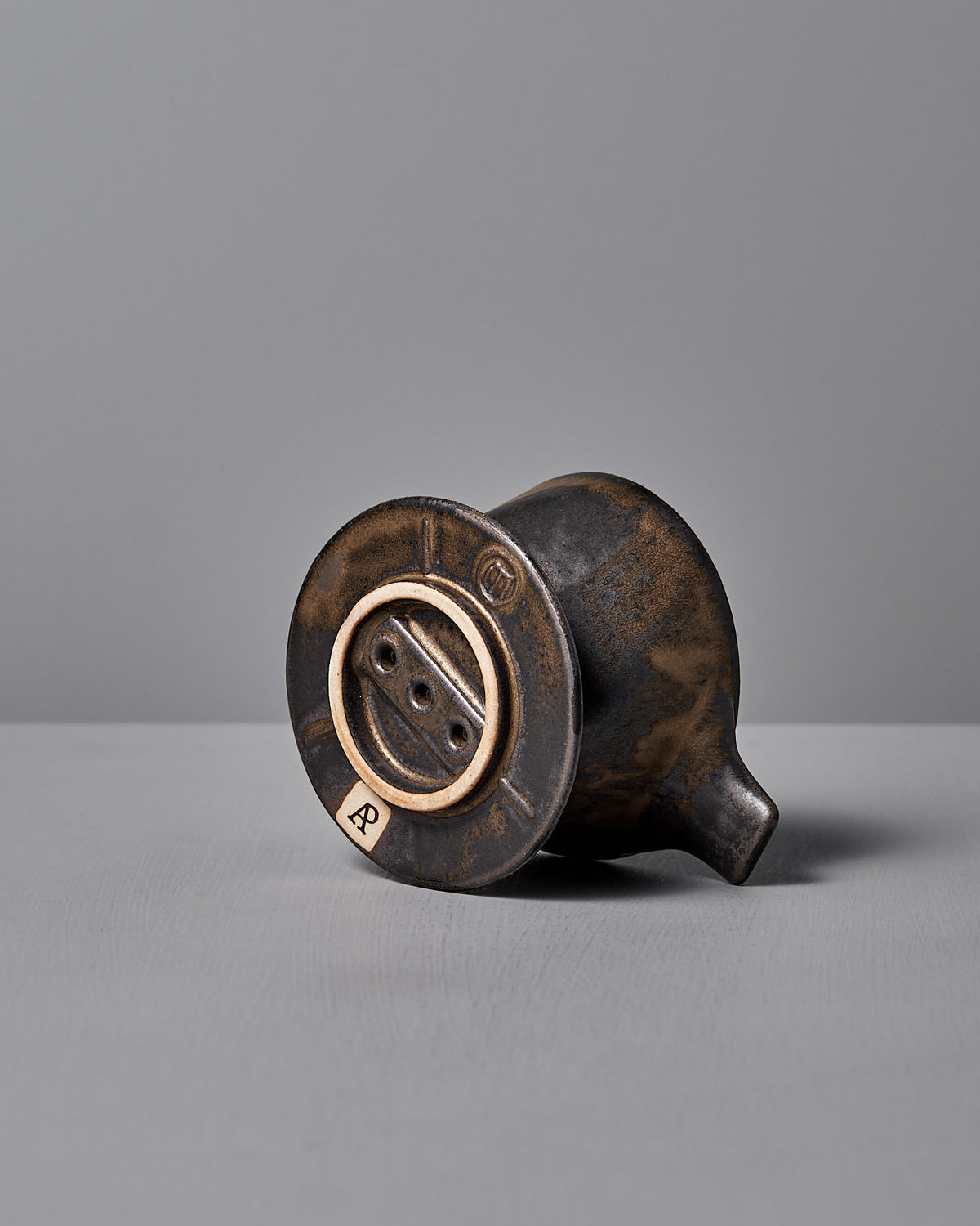 A small, vintage Mino-yaki Coffee Dripper (2 Cup) – Brass by CHIPS Inc. lies on its side revealing its round base and drainage holes against a plain gray background.