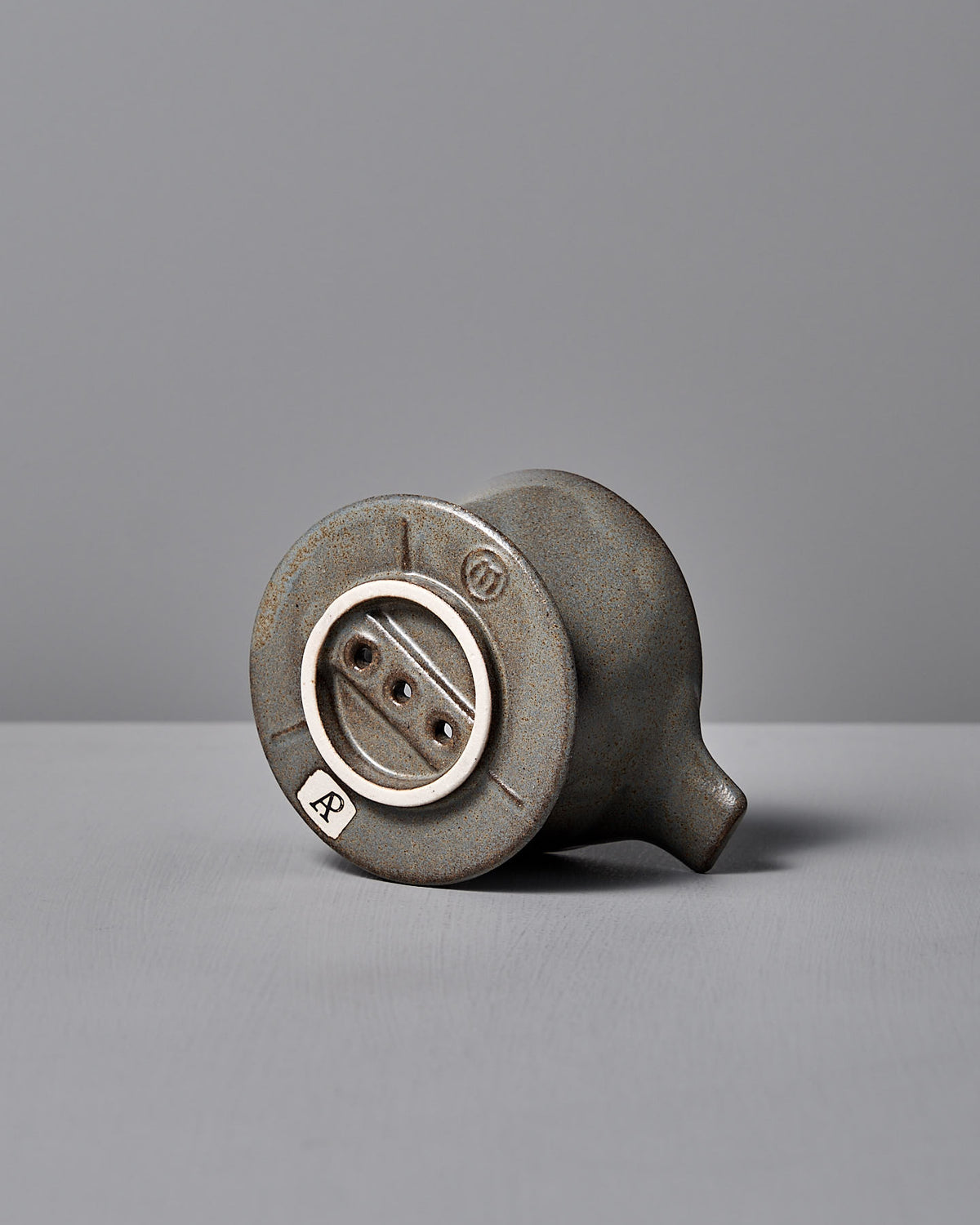 A Mino-yaki Coffee Dripper from CHIPS Inc., resembling a vintage light socket, lies on its side against a gray backdrop, showcasing timeless elegance.