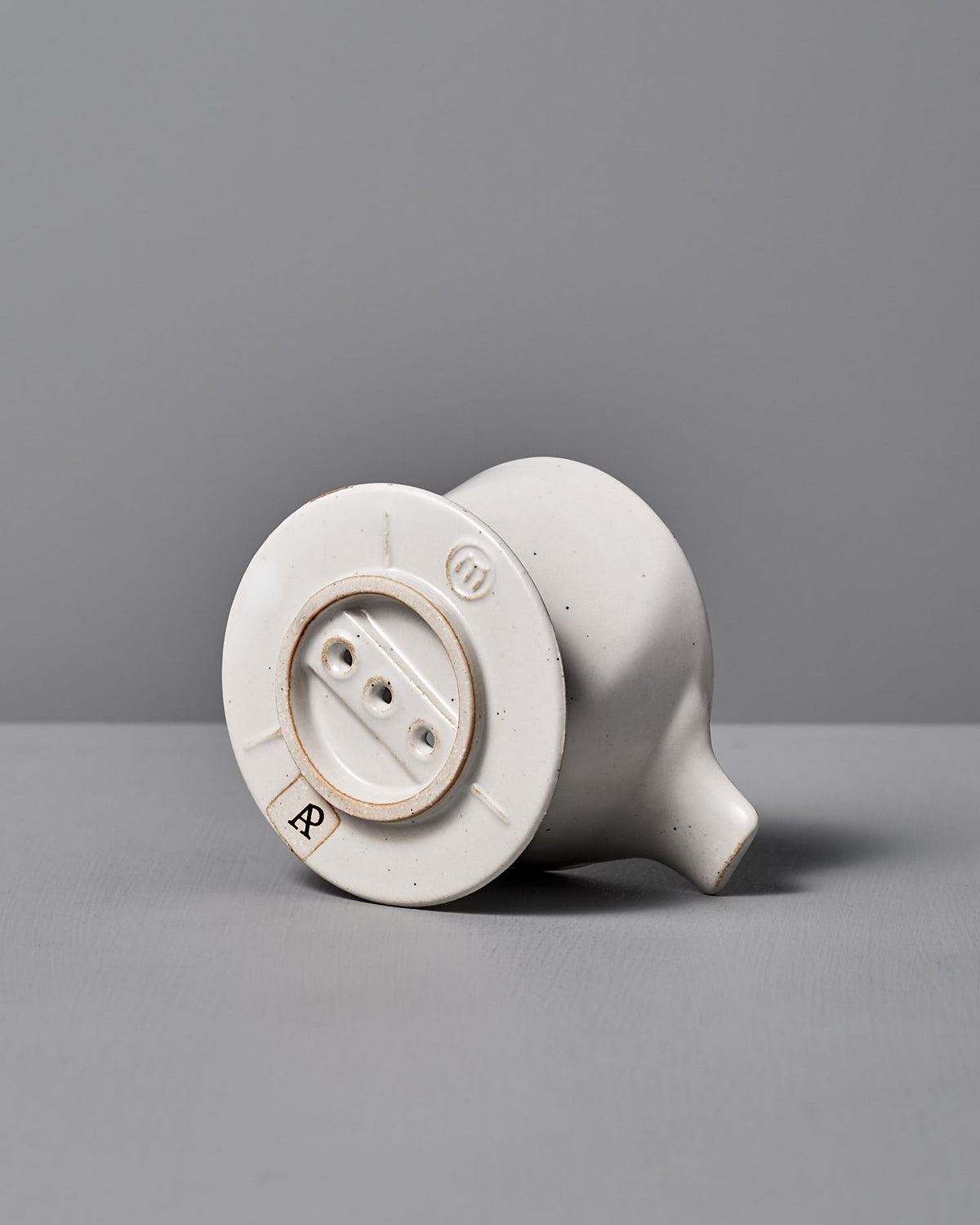 The Mino-yaki Coffee Dripper by CHIPS Inc. features a smooth, rounded ceramic shape with a small opening. Its base, adorned with a textured design and three holes, bears the letter "R," showcasing its origin as an ancient Japanese pottery piece.