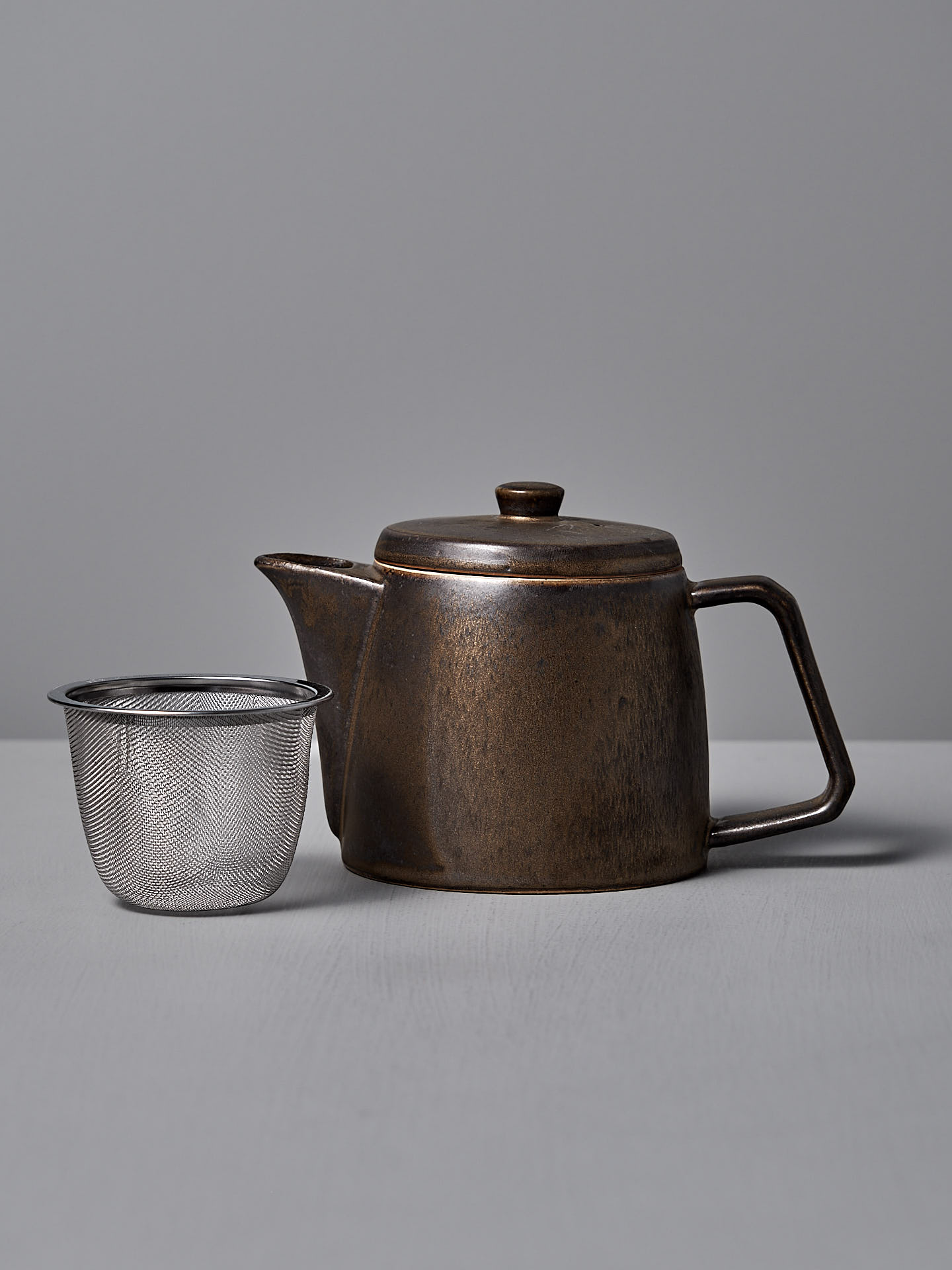 A CHIPS Inc. Mino-yaki Teapot (2 Cup) in dark brown brass with a lid and angular handle sits next to a stainless steel mesh tea infuser on a light gray surface.