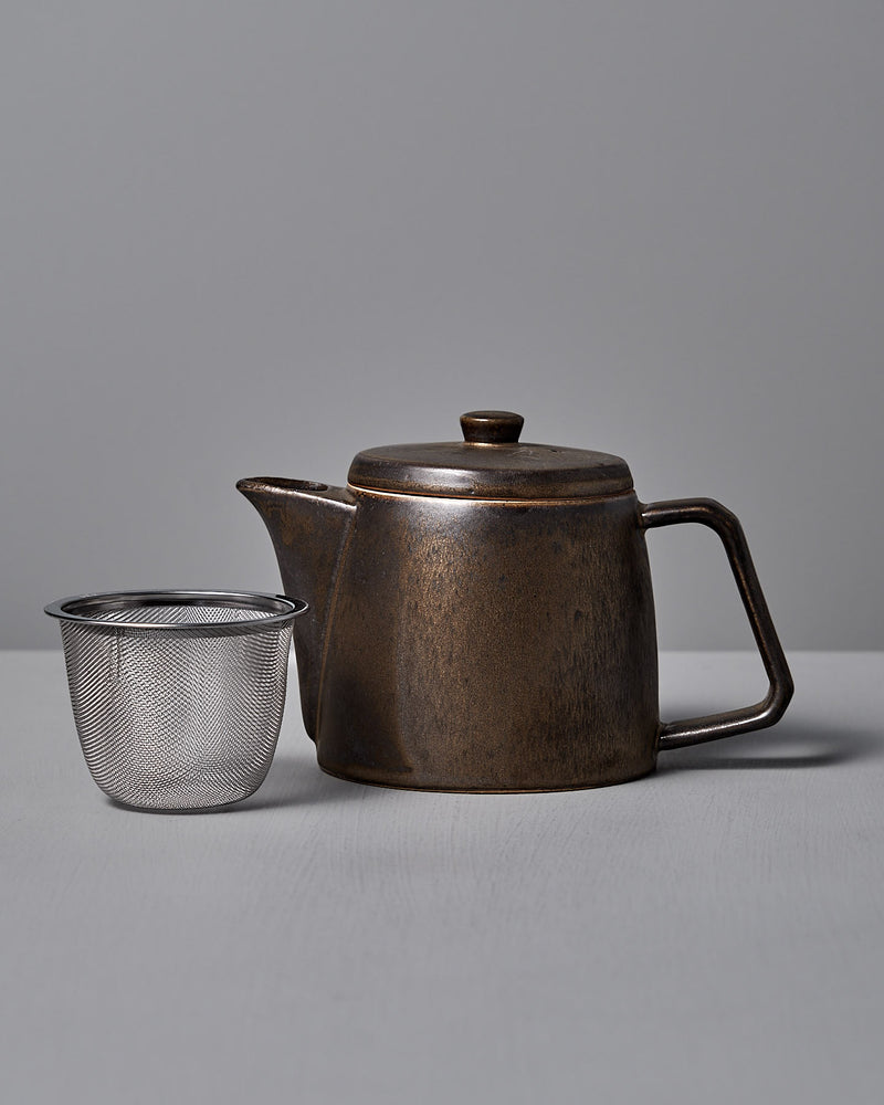 A CHIPS Inc. Mino-yaki Teapot (2 Cup) in dark brown brass with a lid and angular handle sits next to a stainless steel mesh tea infuser on a light gray surface.