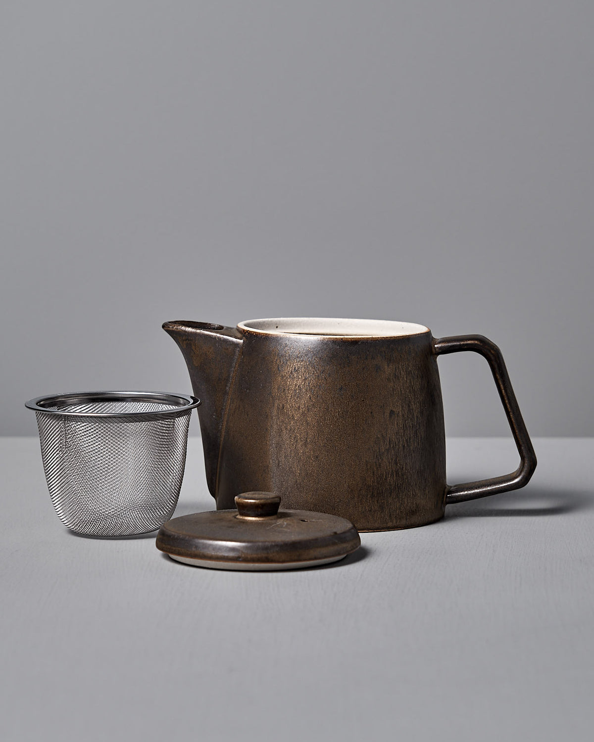The CHIPS Inc. Mino-yaki Teapot (2 Cup) – Brass is a brown ceramic teapot with a matching lid, crafted using the Mino-yaki technique. It includes a separate metal strainer and rests elegantly on the gray surface.