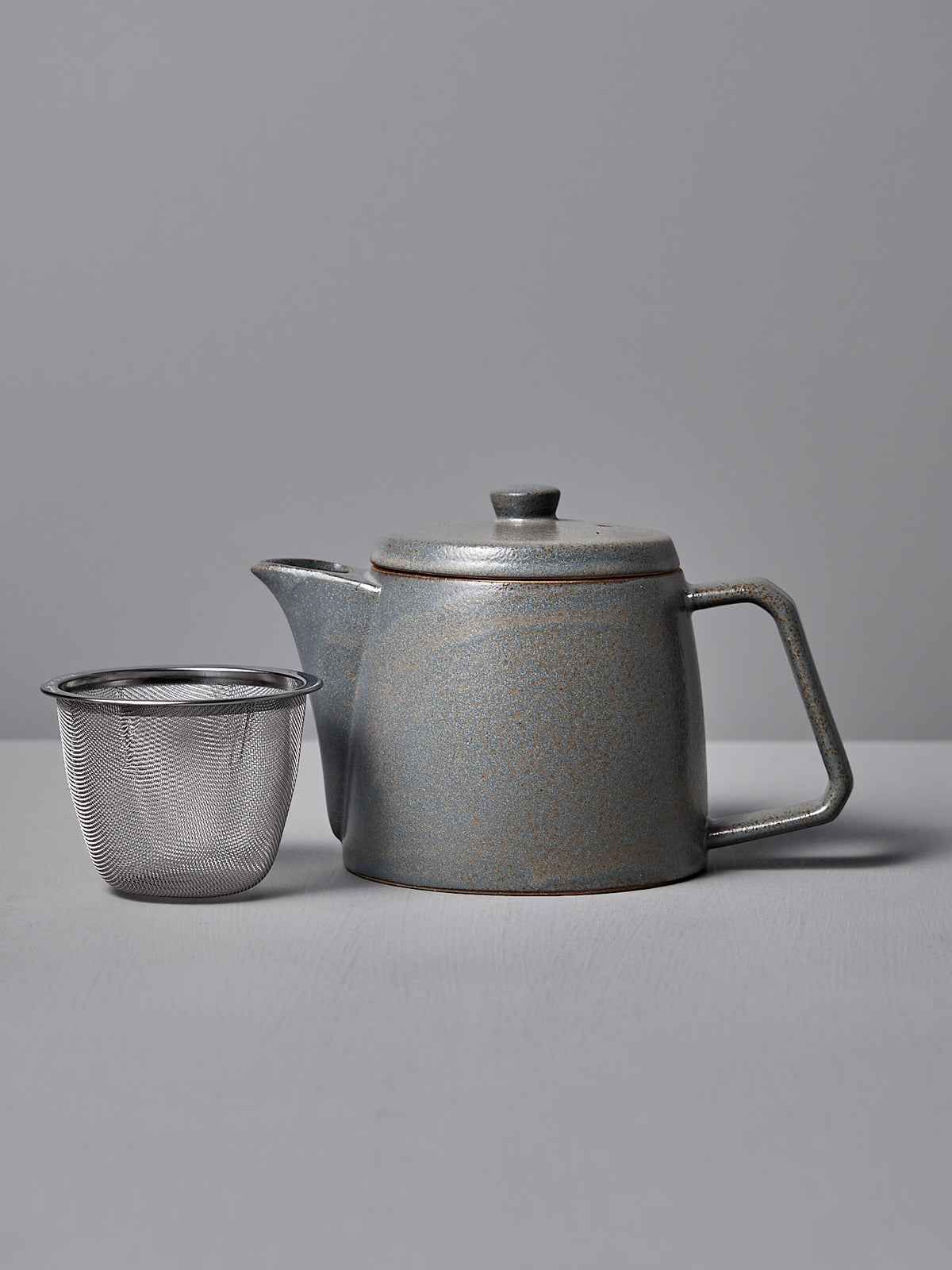 The Mino-yaki Teapot (2 Cup) – Grey by CHIPS Inc., featuring a lid and handle, rests beside a metal mesh infuser, perfect for loose leaf tea, on a gray surface.