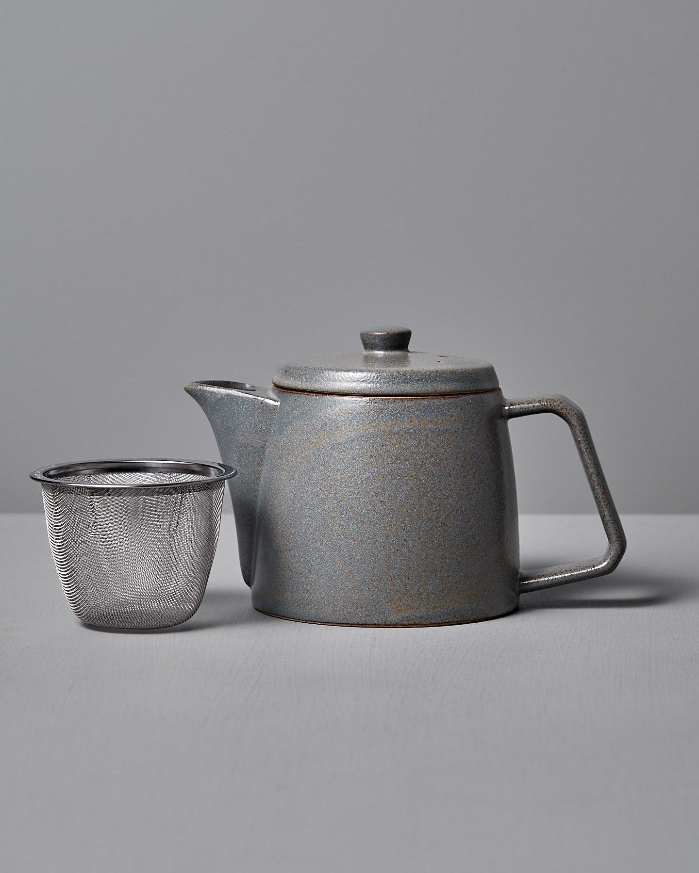The Mino-yaki Teapot (2 Cup) – Grey by CHIPS Inc., featuring a lid and handle, rests beside a metal mesh infuser, perfect for loose leaf tea, on a gray surface.