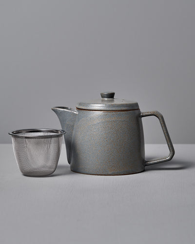 The Mino-yaki Teapot (2 Cup) – Grey by CHIPS Inc., featuring a lid and handle, rests beside a metal mesh infuser, perfect for loose leaf tea, on a gray surface.