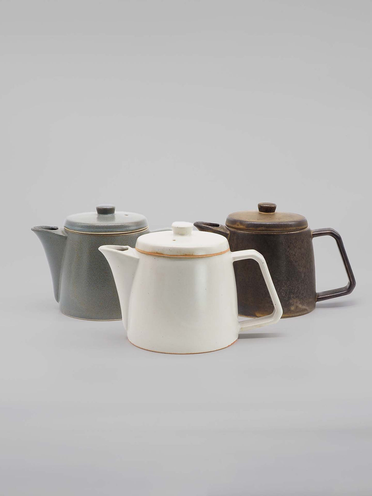 Three Mino-yaki teapots by CHIPS Inc., crafted in varying colors of white, dark gray, and brown, are artfully arranged on a neutral background.