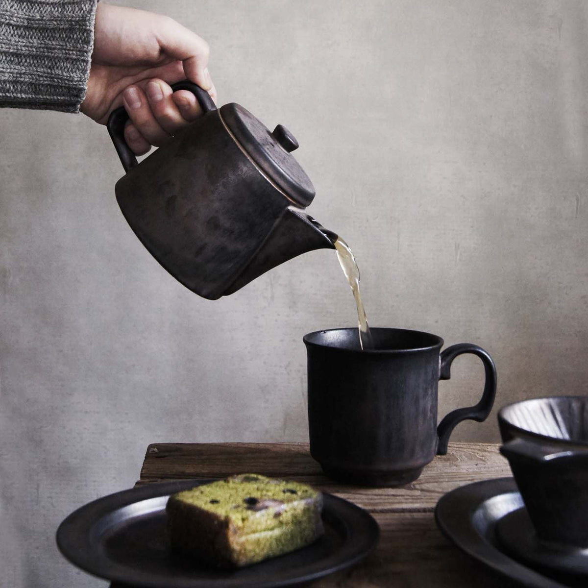 A hand pours loose leaf tea from a CHIPS Inc. Mino-yaki Teapot (2 Cup) – Brass into a matching cup on a wooden table. A slice of green bread rests on a plate beside the teapot.