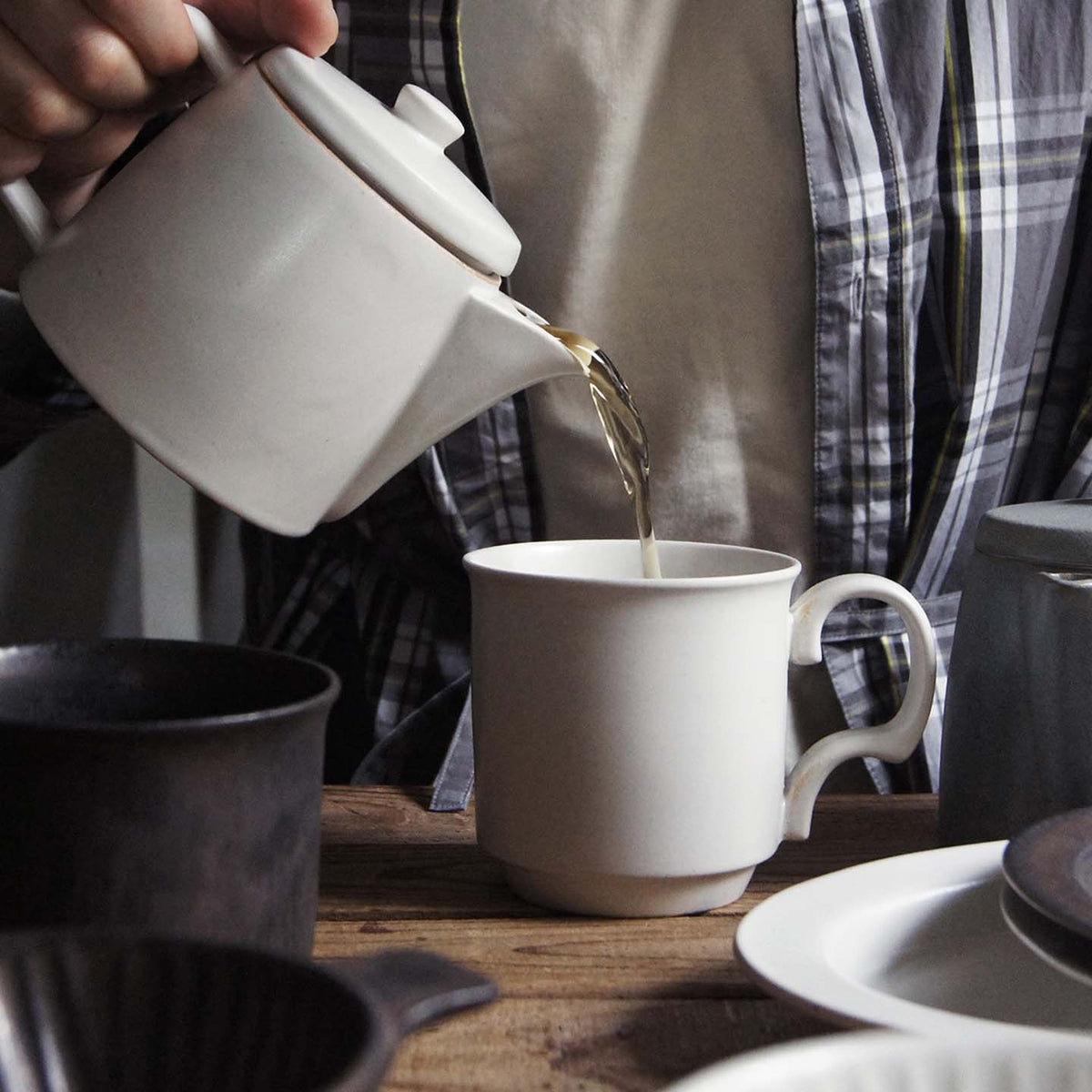 A person pours loose leaf tea from the CHIPS Inc. Mino-yaki Teapot (2 Cup) – White, crafted using the traditional technique, into a white mug on a wooden table adorned with cups and plates.