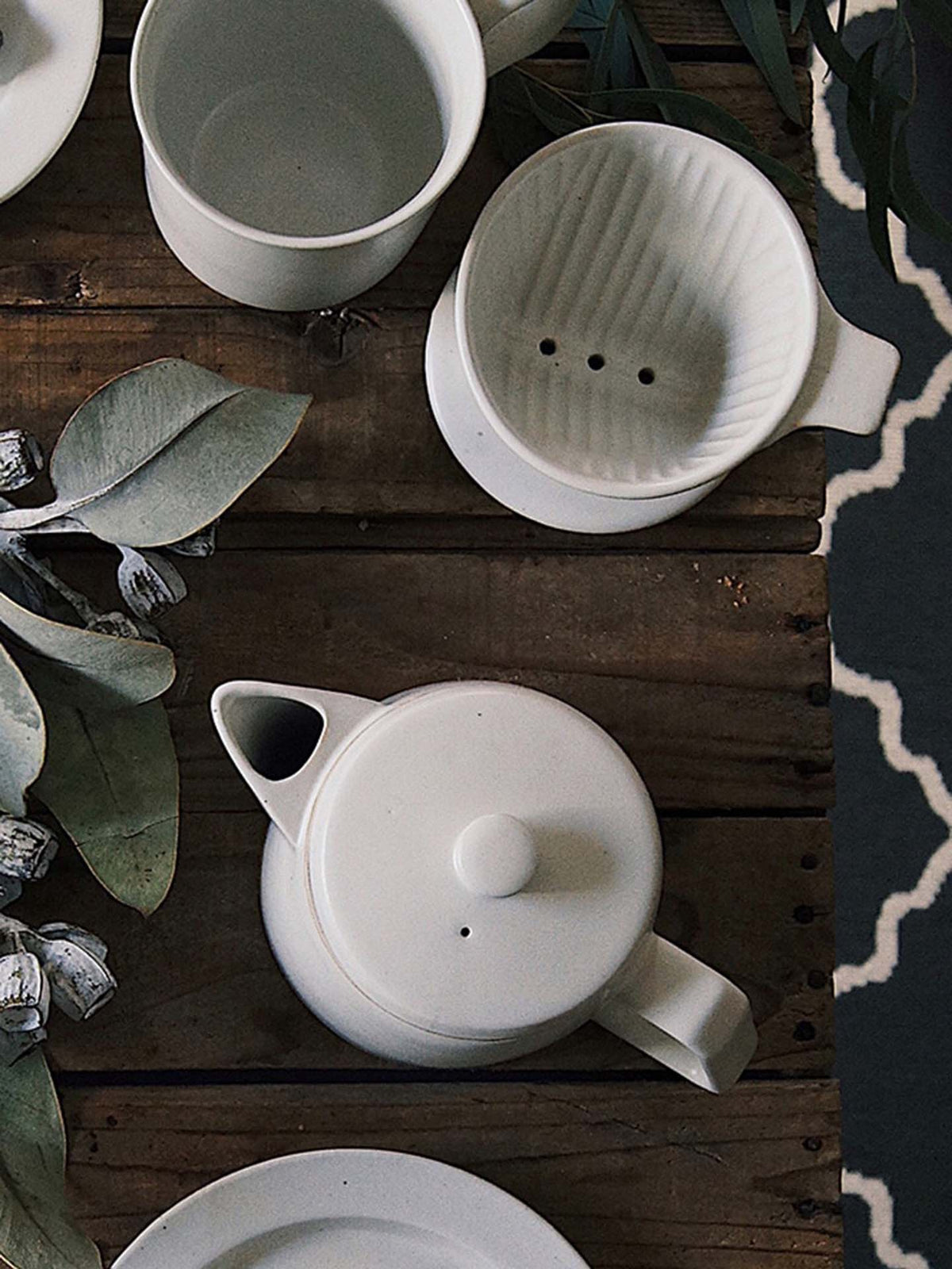 White Mino-yaki Coffee Dripper (2 Cup) by CHIPS Inc. accompanied by a ceramic teapot and cup, resting on a wooden table surrounded by eucalyptus leaves.