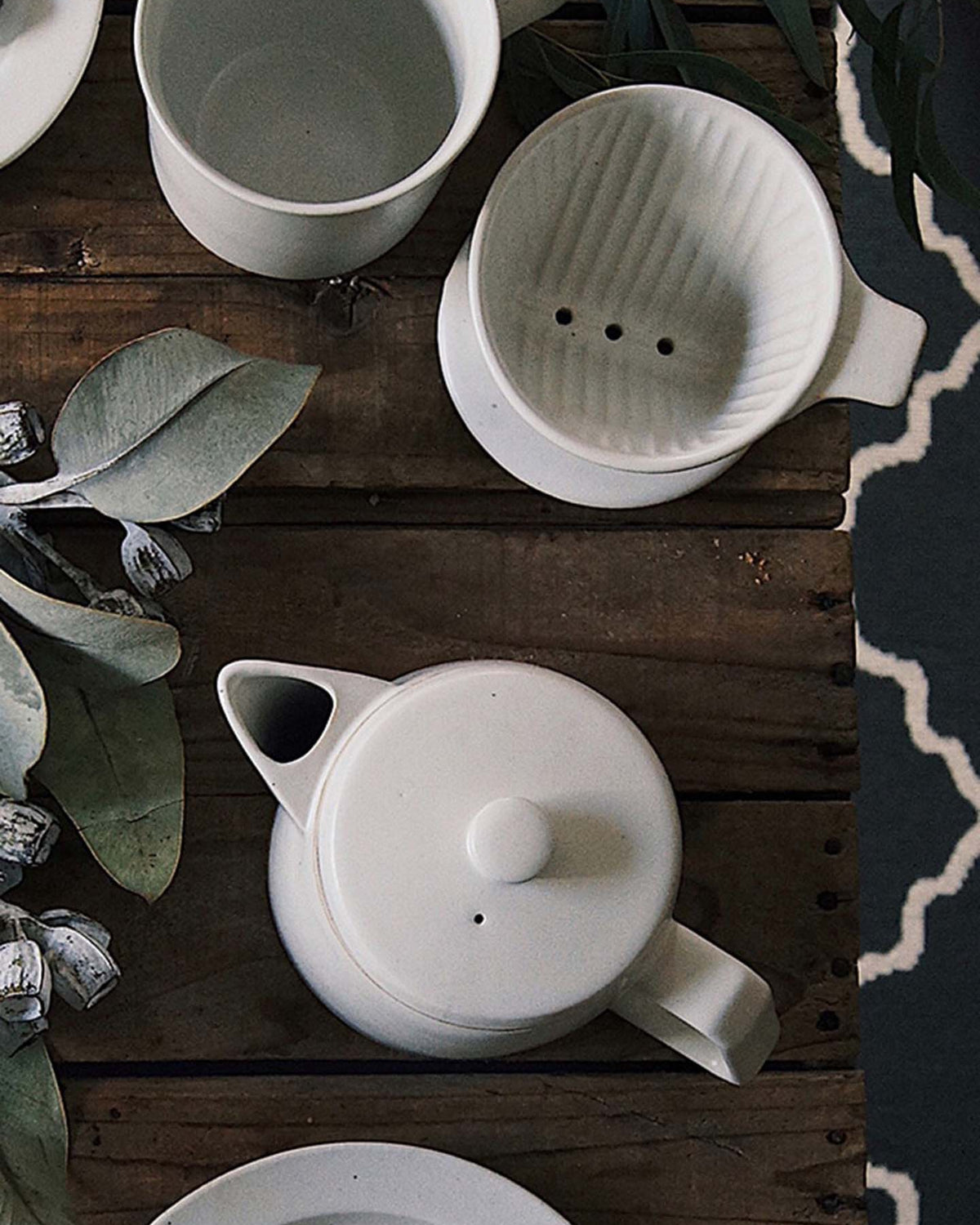 White Mino-yaki Coffee Dripper (2 Cup) by CHIPS Inc. accompanied by a ceramic teapot and cup, resting on a wooden table surrounded by eucalyptus leaves.