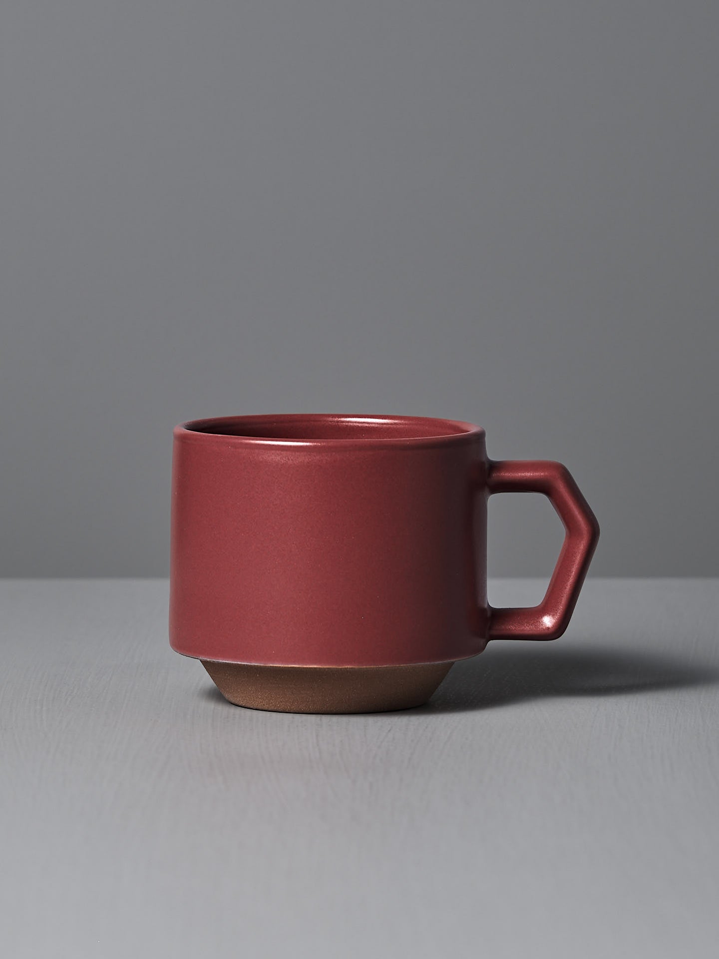 The Stacking Mug – Matte Red by CHIPS Inc., styled with a sleek angular handle and reminiscent of elegant Minoyaki porcelain, sits gracefully on a gray surface.