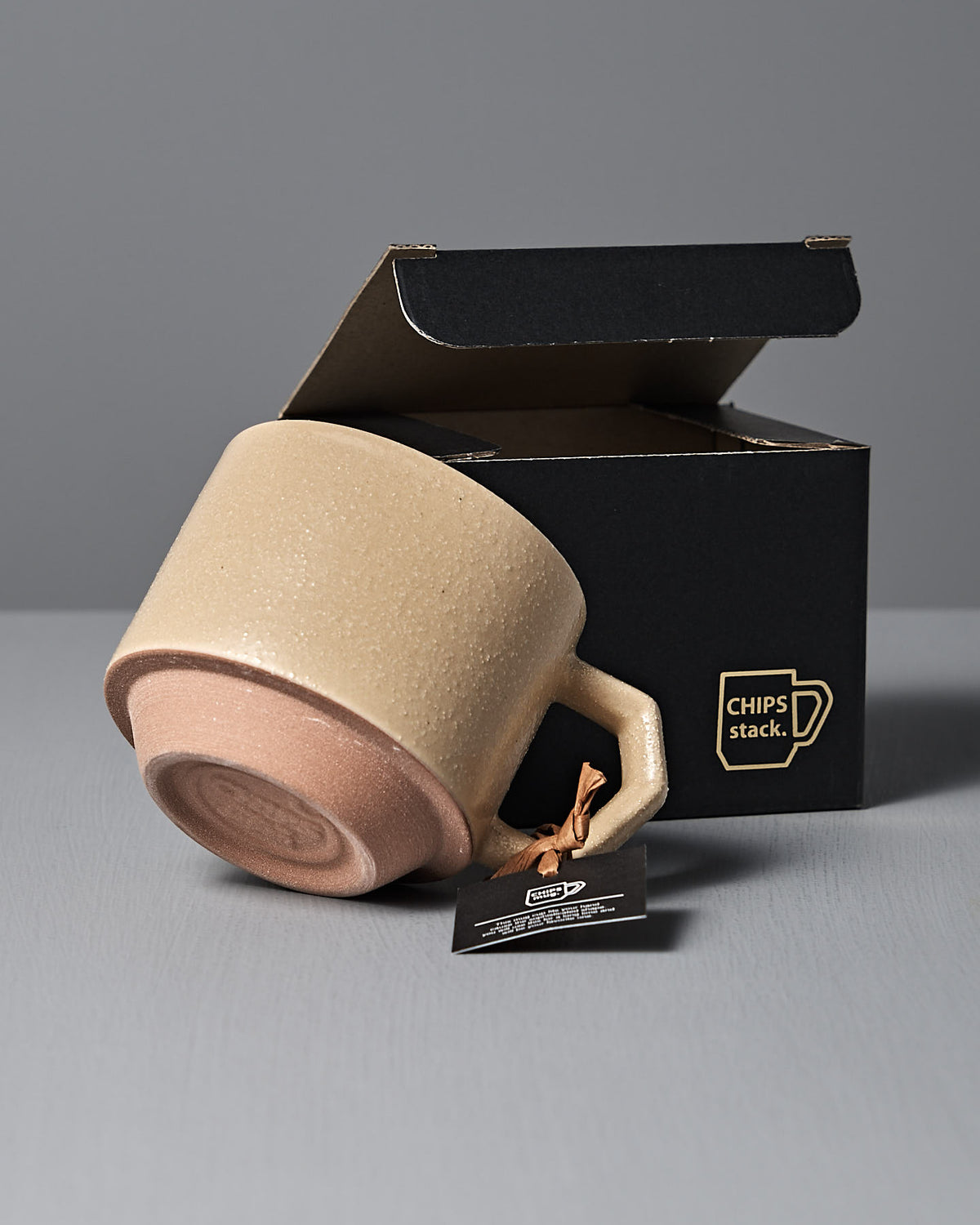 A beautifully crafted ceramic mug in sand tan, made from Japanese porcelain, sits partially outside a black "CHIPS stack" box by CHIPS Inc., with a small tag on the handle.