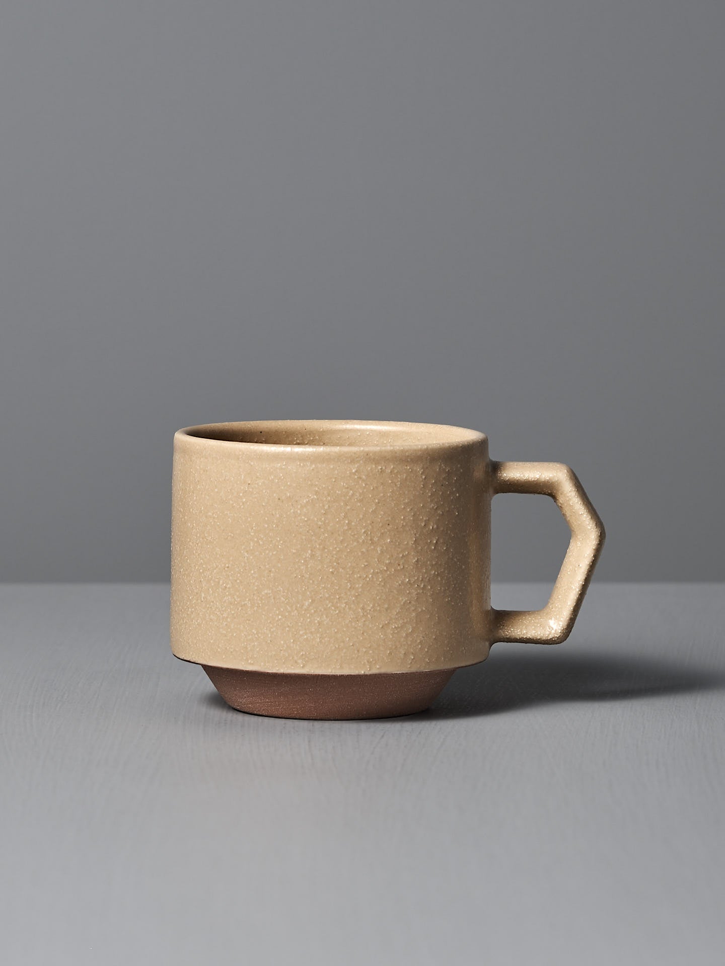 A CHIPS Inc. Stacking Mug – Sand Tan sits on a gray surface against a gray background, highlighting the elegance of Japanese porcelain with its geometric handle.
