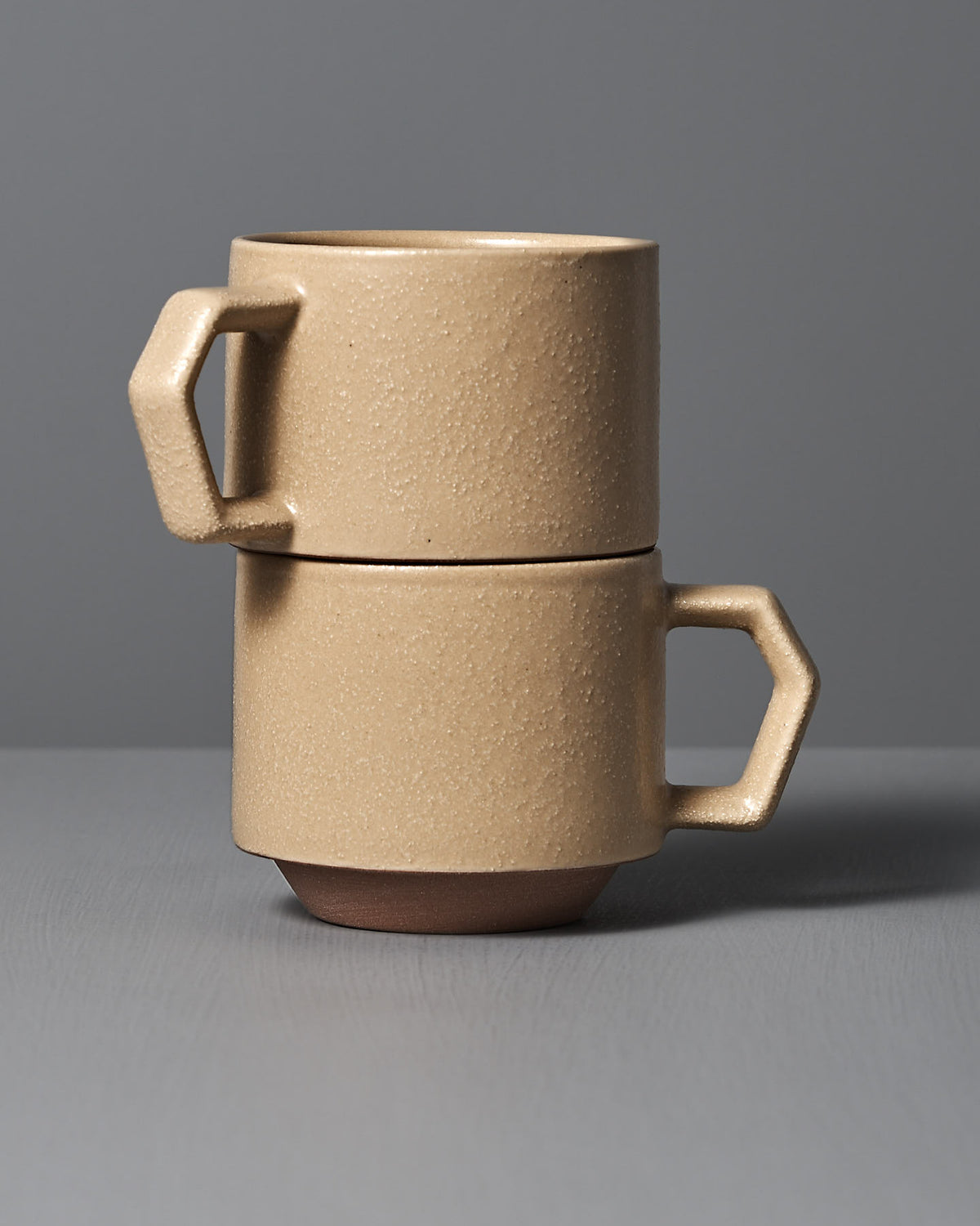 The Stacking Mug – Sand Tan by CHIPS Inc., inspired by Japanese porcelain, features two stackable, beige ceramic mugs with angular handles resting elegantly on a gray surface.