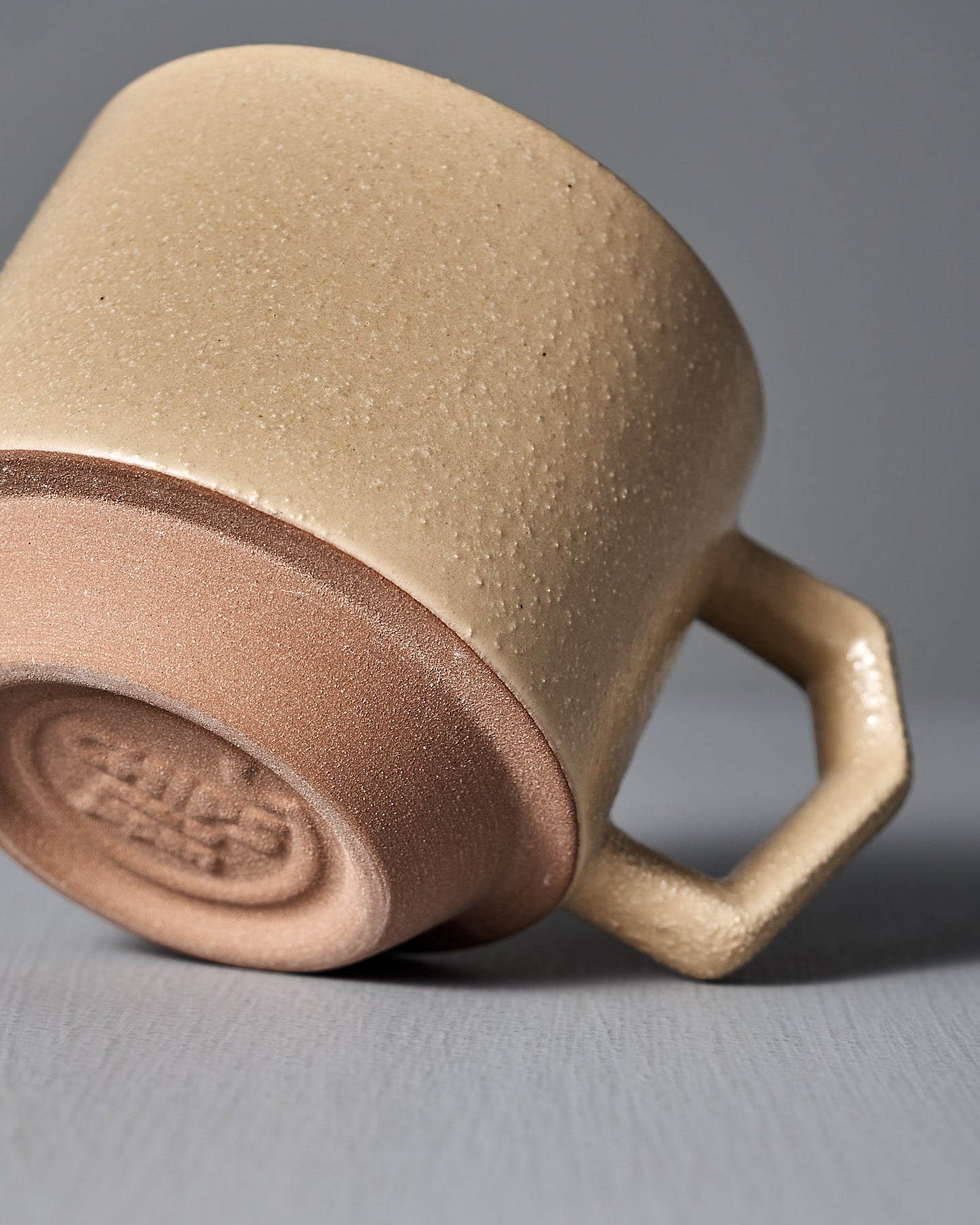 A CHIPS Inc. Stacking Mug – Sand Tan sits on a gray surface against a gray background, highlighting the elegance of Japanese porcelain with its geometric handle.