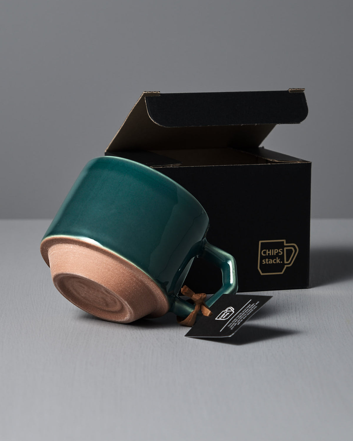 A Stacking Mug – Teal from CHIPS Inc. rests on its side with a cardboard box in the background, reflecting the brand's elegance.
