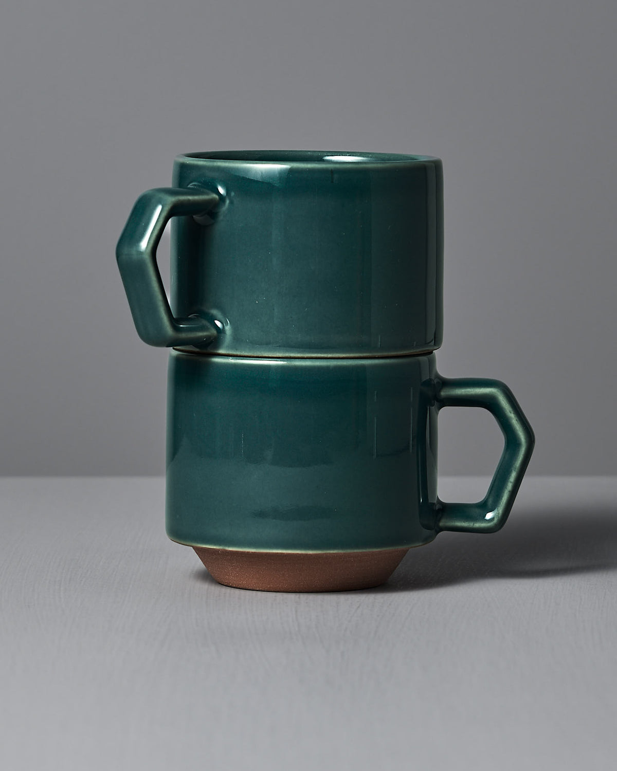 Two teal stacking mugs by CHIPS Inc., featuring hexagonal handles, are displayed on a gray surface. Part of the CHIPS tableware collection, they elegantly enhance any setting.