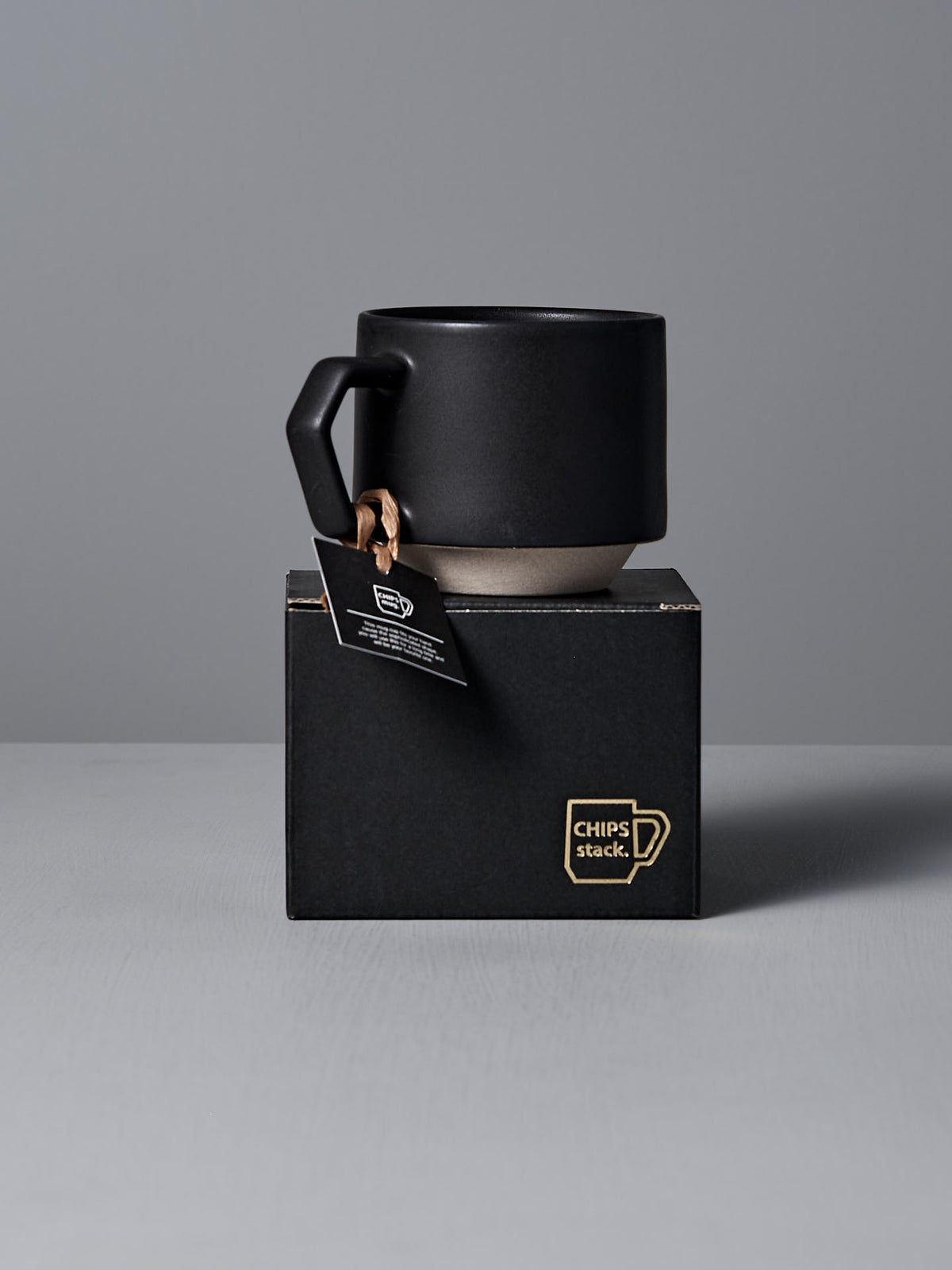 A sophisticated Stacking Mug – Black from CHIPS Inc. features a textured handle and rests on a branded black box labeled &quot;CHIPS Stack,&quot; with its stackable design accentuated by a tag affixed to the handle.