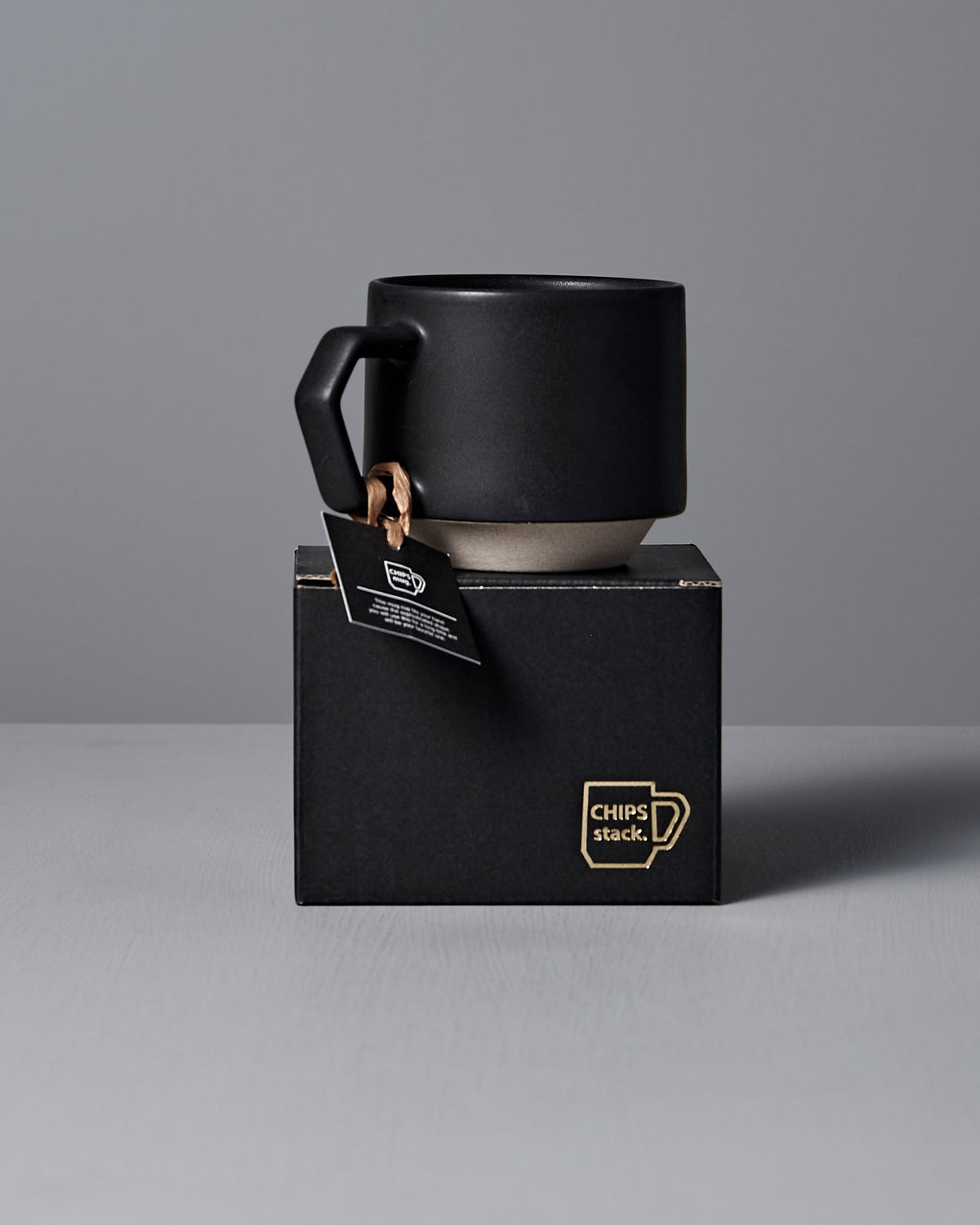 A black Stacking Mug – Matte Black with a square handle from CHIPS Inc. sits on a black box labeled "CHIPS stack," with a tag hanging from the handle.