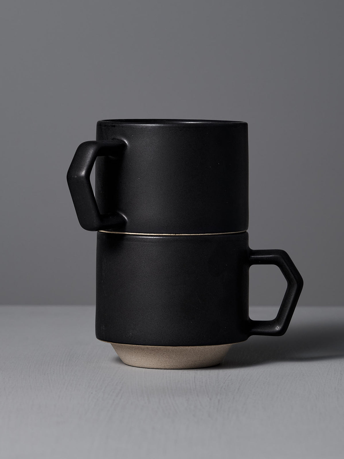 Two Stacking Mug – Black pieces by CHIPS Inc. crafted from Minoyaki porcelain, featuring geometric handles, are elegantly arranged on a gray surface against a plain gray background.