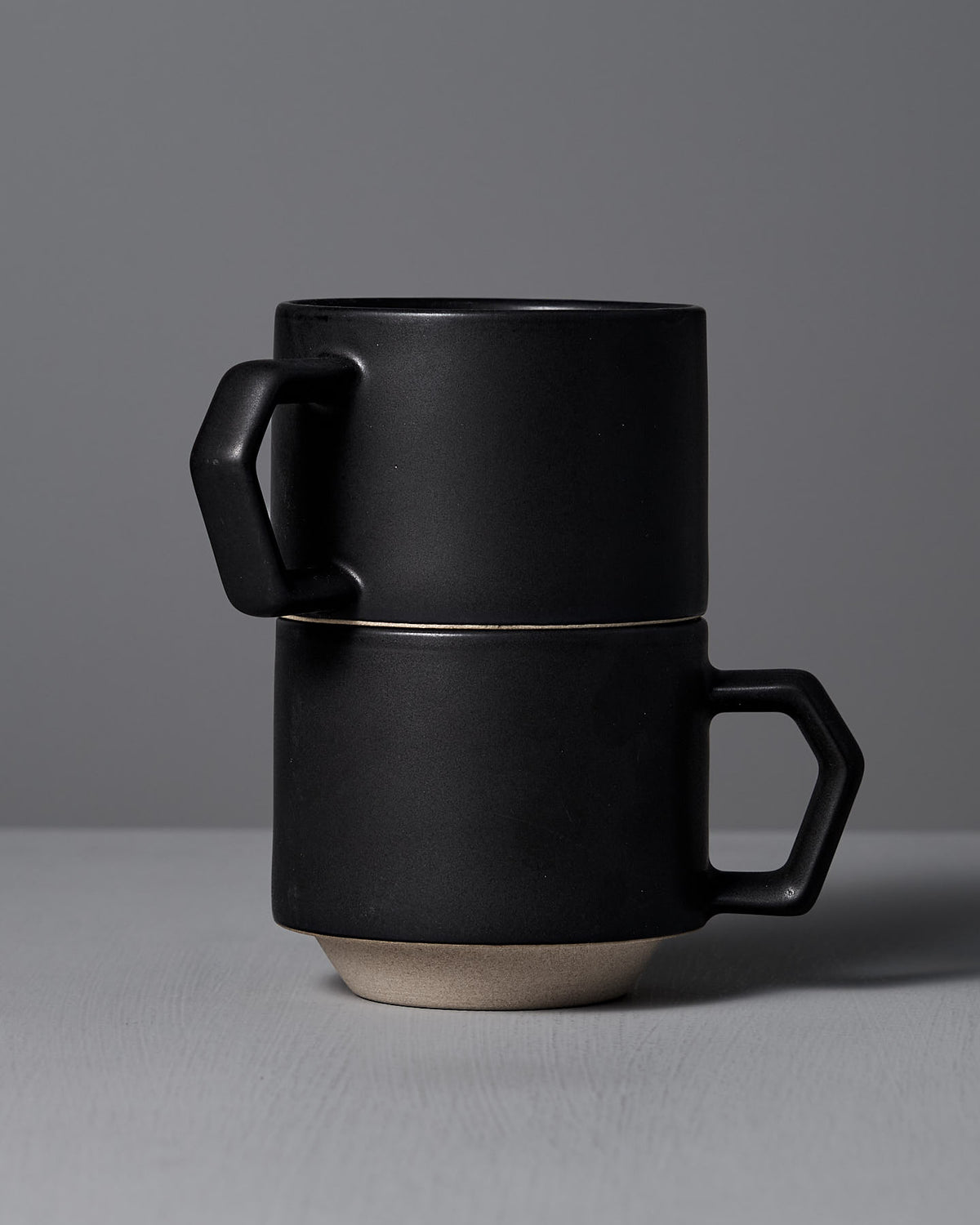Two Stacking Mugs in Matte Black by CHIPS Inc. with angular handles rest against a gray background, highlighting their artisanal elegance.