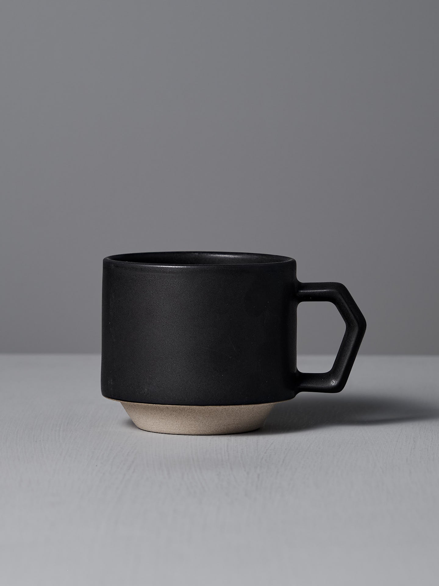 A CHIPS Inc. Stacking Mug in matte black, featuring a geometric handle, rests elegantly on a light gray surface against a gray background.