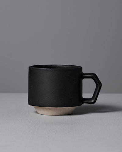 A CHIPS Inc. Stacking Mug in matte black, featuring a geometric handle, rests elegantly on a light gray surface against a gray background.