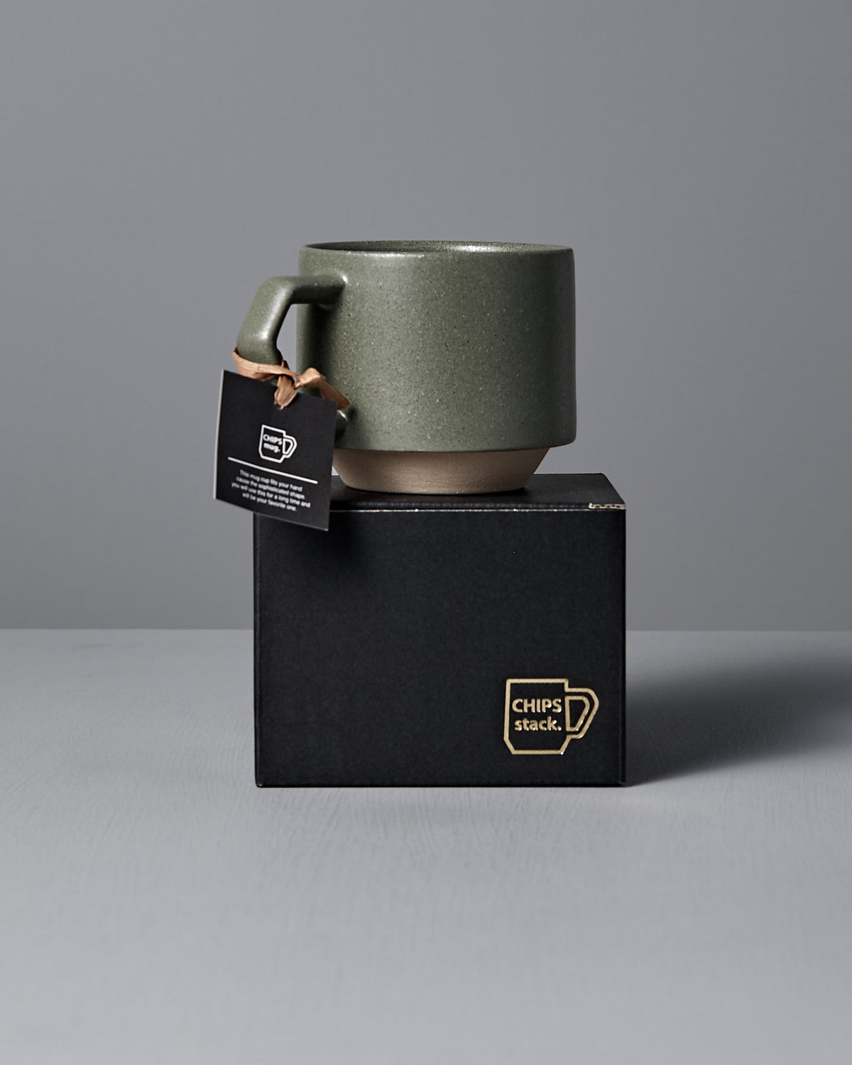 A CHIPS Inc. Stacking Mug in sand green, complete with a tag, rests elegantly atop a sleek black box.