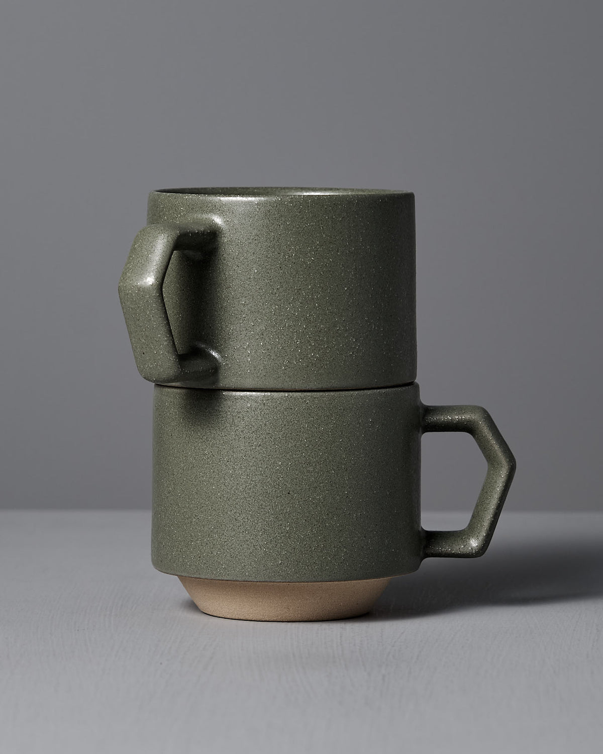 Two CHIPS Inc. Stacking Mugs in Sand Green with angular handles, crafted from Minoyaki porcelain, are stacked on a gray surface.