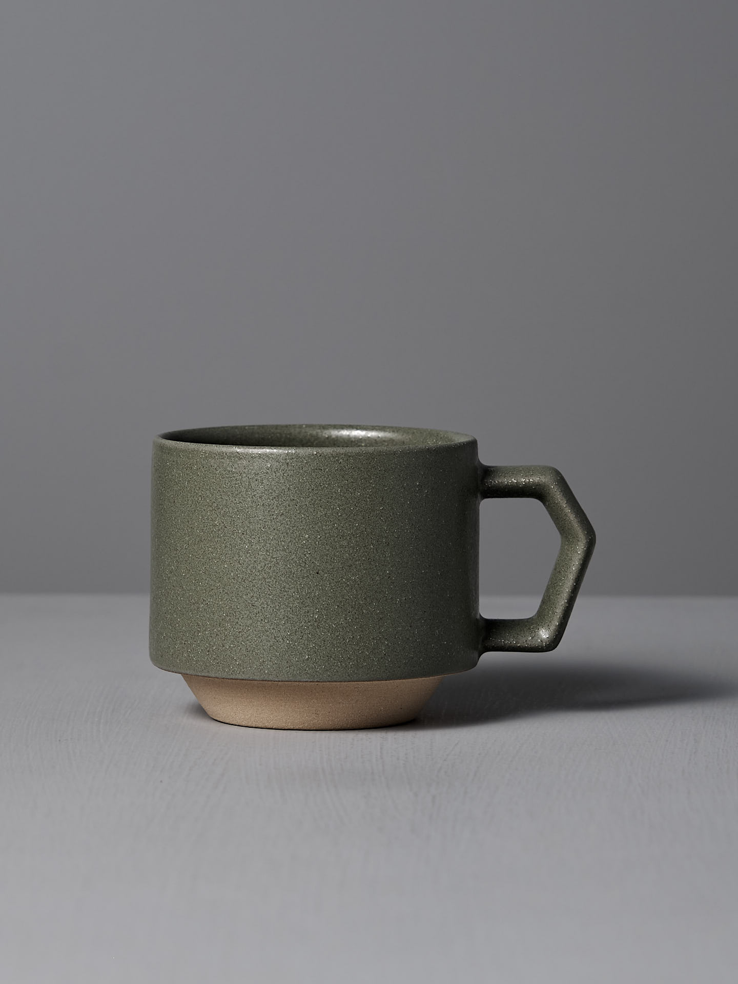 The CHIPS Inc. Stacking Mug – Sand Green, with a geometric handle and resembling Minoyaki porcelain, is displayed on a gray surface against a gray backdrop.