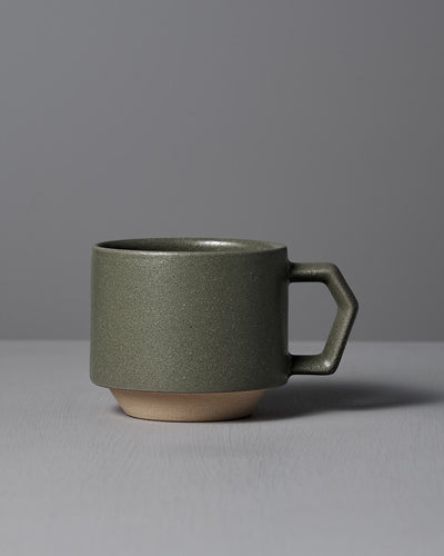 The CHIPS Inc. Stacking Mug – Sand Green, with a geometric handle and resembling Minoyaki porcelain, is displayed on a gray surface against a gray backdrop.