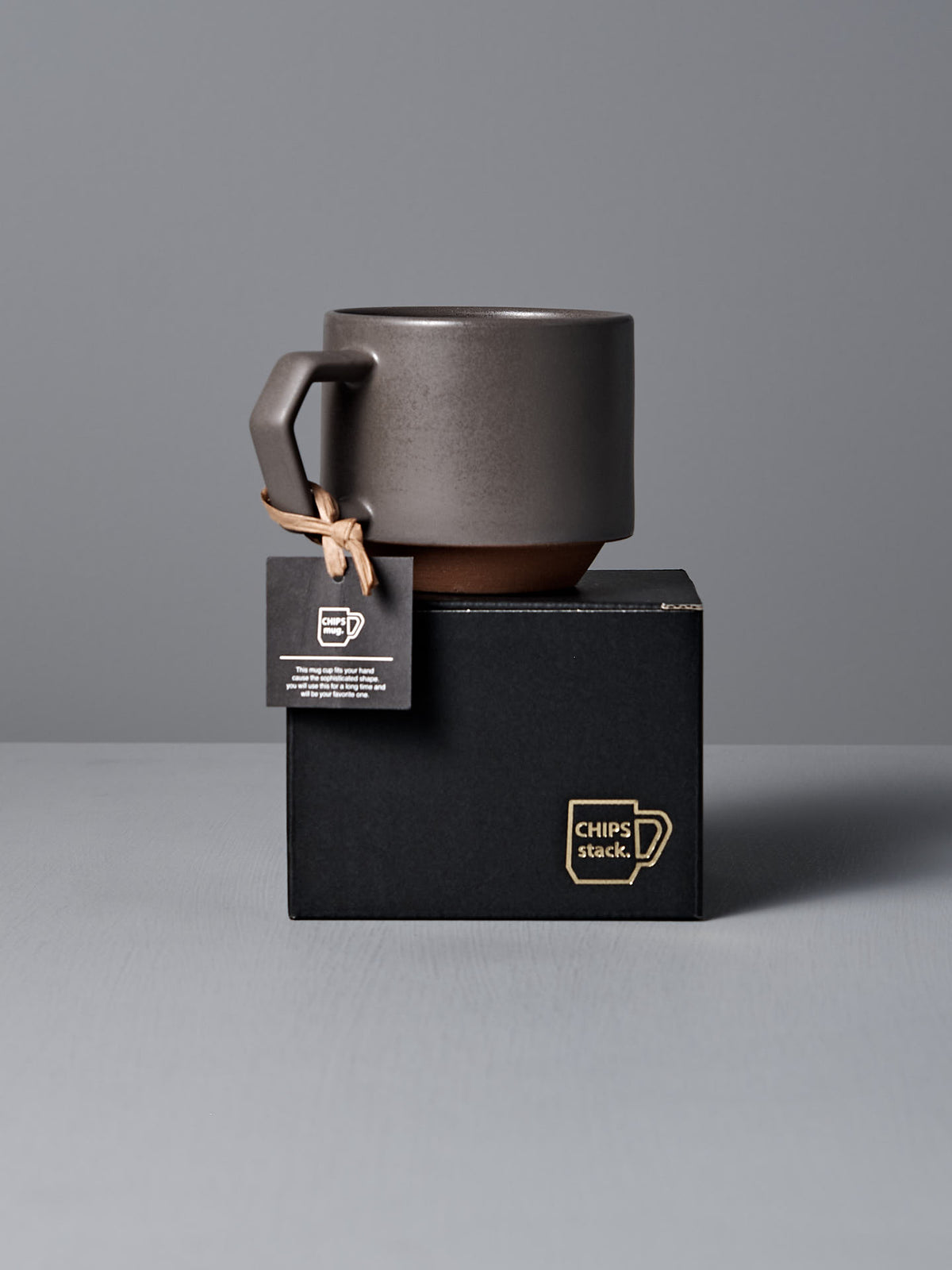 A &quot;Stacking Mug – Grey&quot; by CHIPS Inc., made from Minoyaki porcelain with an angular handle, rests on a black box marked &quot;CHIPS stack,&quot; with a tag attached to the handle.