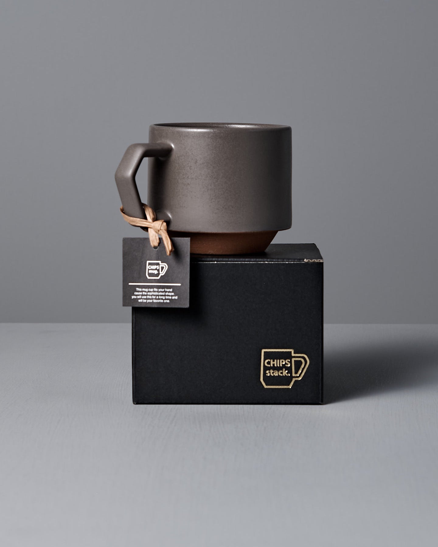 The Stacking Mug – Matte Grey by CHIPS Inc. is a sleek, dark ceramic piece featuring a geometric handle, elegantly resting on a gray surface against a matching background.