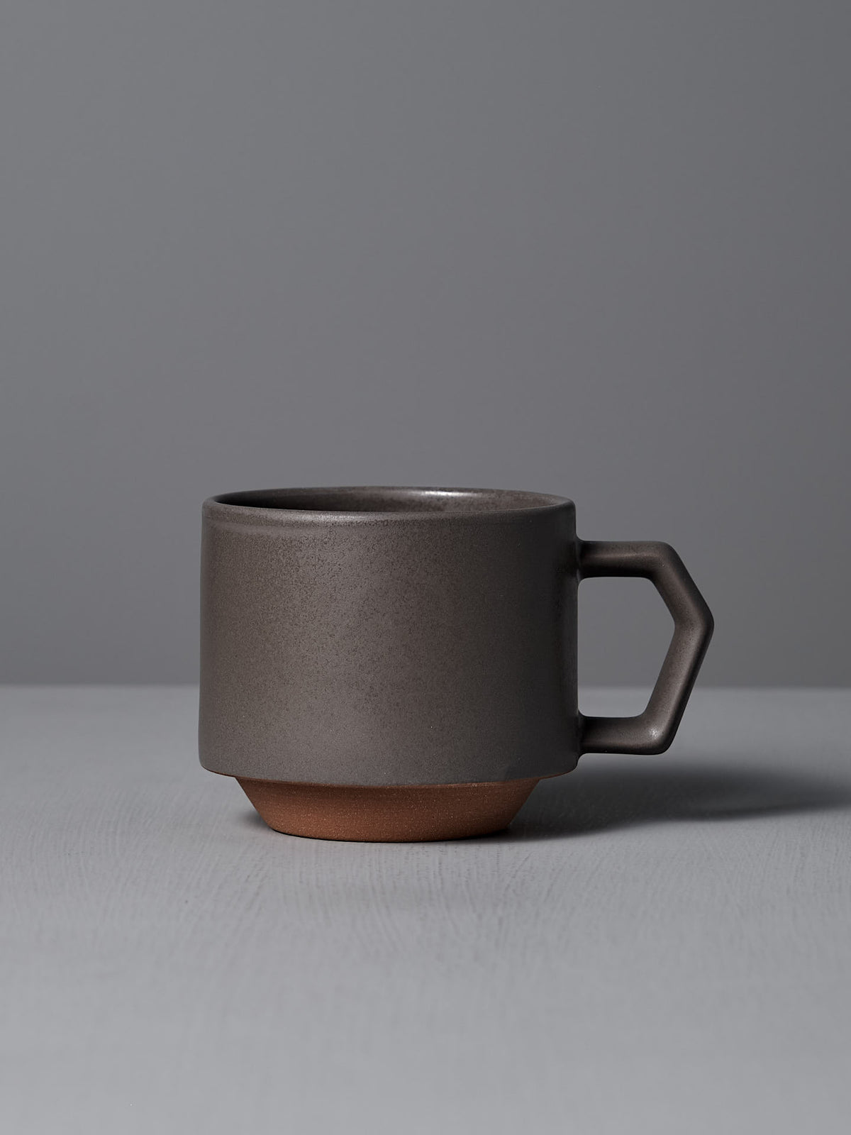 The Stacking Mug – Grey by CHIPS Inc., boasting a unique geometric handle and a brown base, rests stylishly on a gray surface.