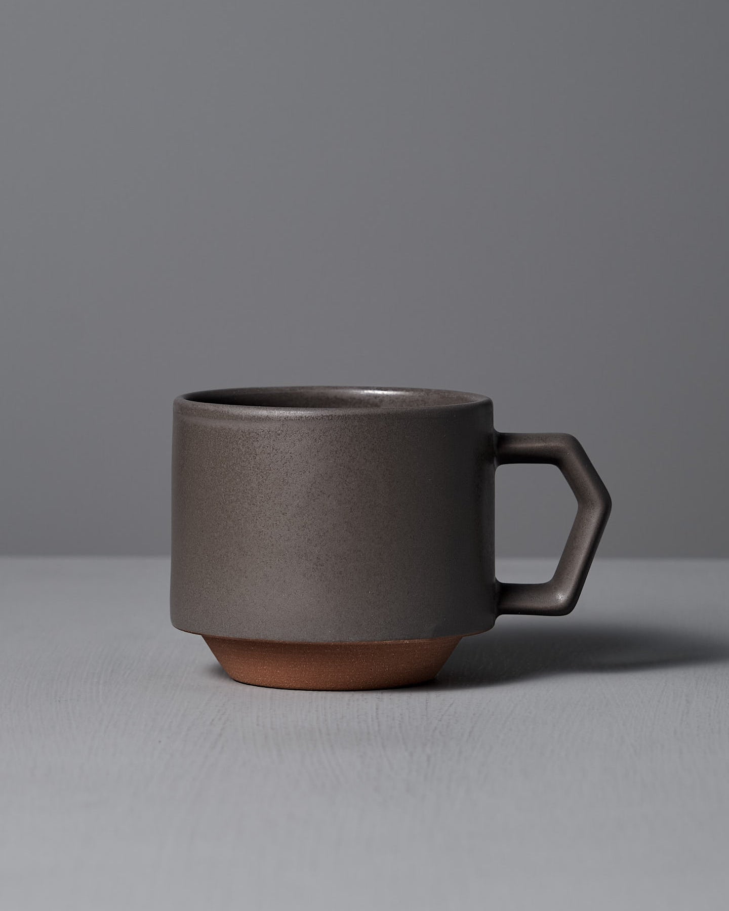 The Stacking Mug – Matte Grey by CHIPS Inc. is a sleek, dark ceramic piece featuring a geometric handle, elegantly resting on a gray surface against a matching background.