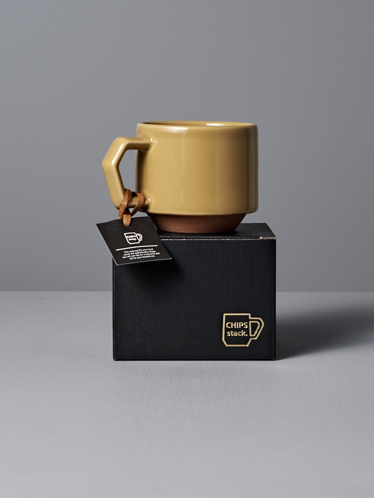 The sophisticated Stacking Mug – Mustard, crafted from Minoyaki porcelain, showcases an angular handle and is elegantly presented on a black box labeled &quot;CHIPS stack&quot; by CHIPS Inc. A tag adorns the mug, set against a simple gray surface and wall that enhances its refined appeal.