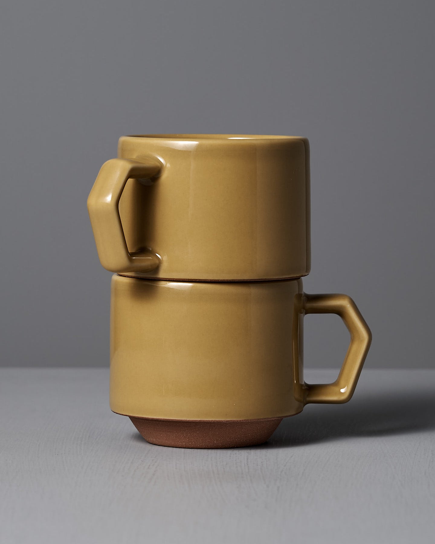 The Stacking Mug – Mustard by CHIPS Inc. exudes elegance as it rests gracefully on a gray surface against a gray background, its geometric handle and matte finish reflecting the refined charm of Minoyaki porcelain.