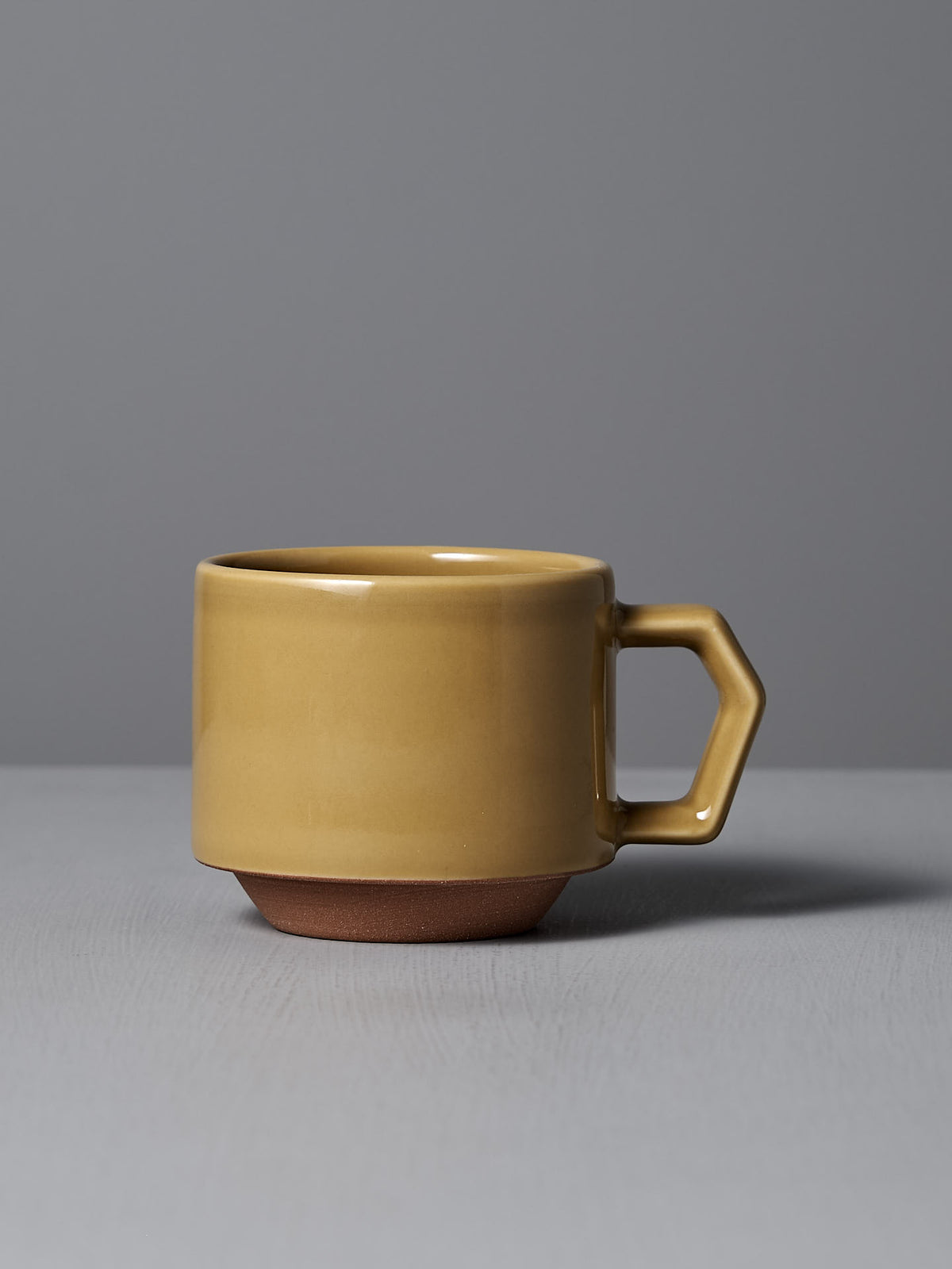 The Stacking Mug – Mustard by CHIPS Inc. exudes elegance as it rests gracefully on a gray surface against a gray background, its geometric handle and matte finish reflecting the refined charm of Minoyaki porcelain.