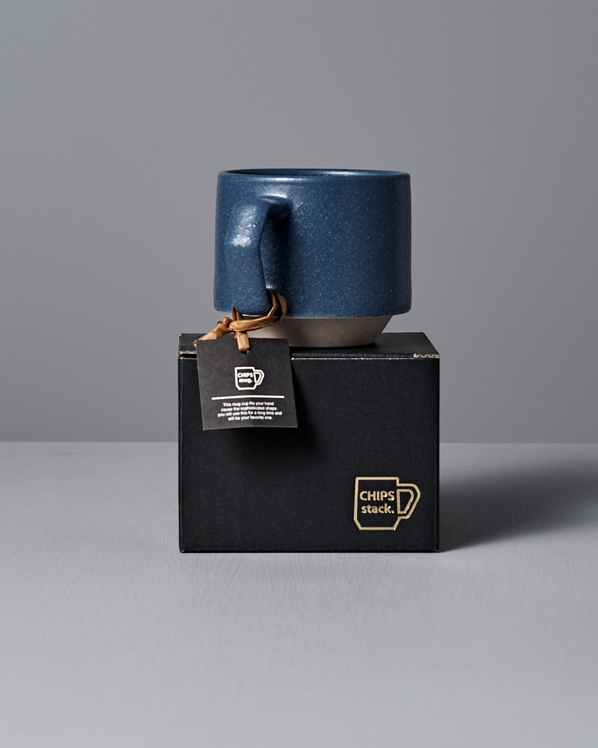A sand blue Stacking Mug from CHIPS Inc. with a tag highlighting its elegant, handmade porcelain design rests atop a black box labeled "CHIPS stack.