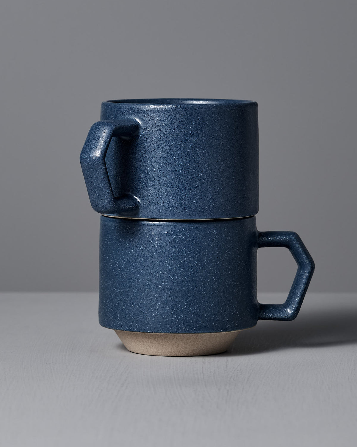 Two stacked Stacking Mugs in Sand Blue by CHIPS Inc. rest on a gray surface against a gray backdrop, highlighting their stackable design and the artistry of handmade porcelain with elegant angular handles.