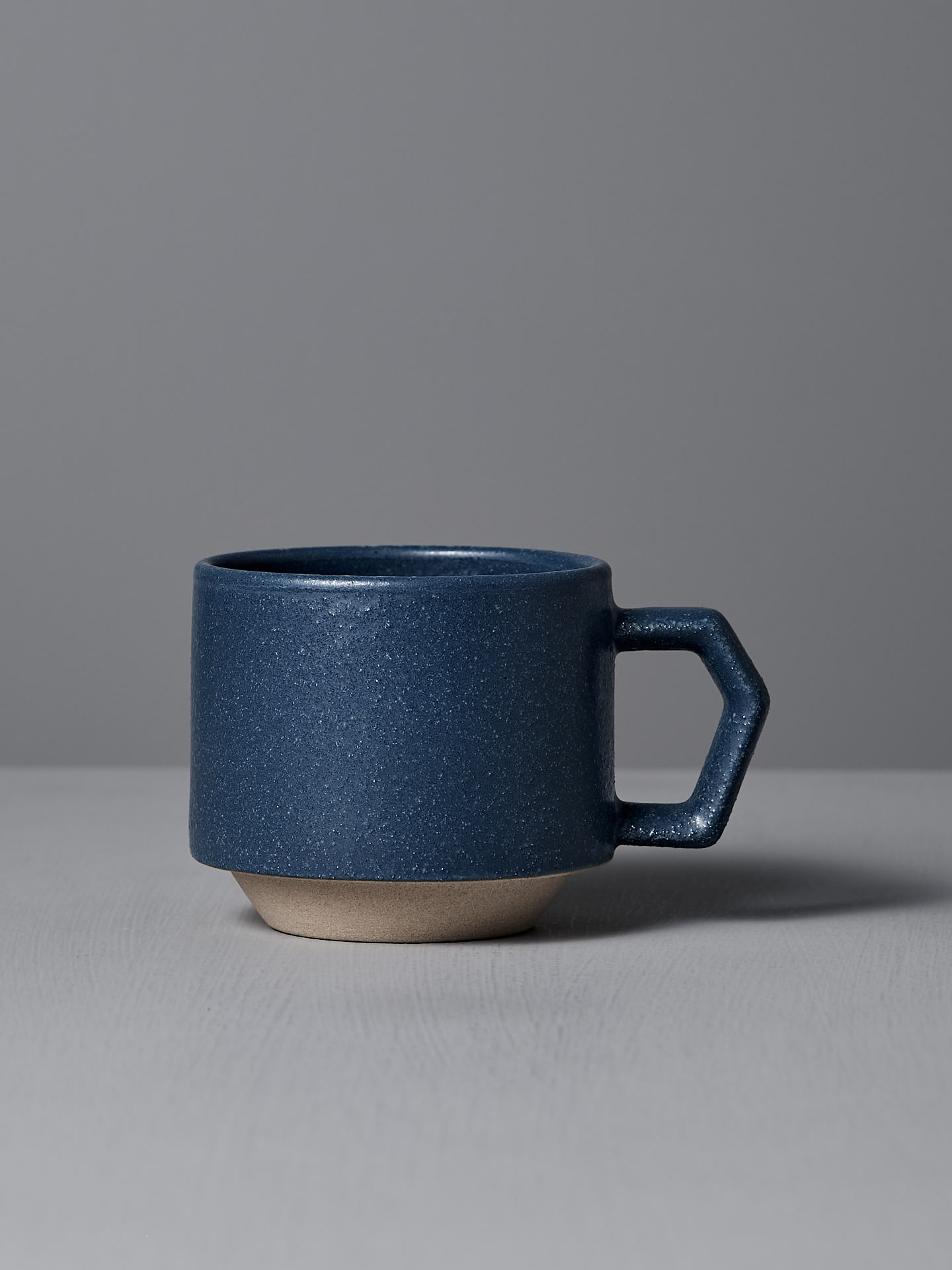 A CHIPS Inc. Stacking Mug in Navy, featuring an angular handle, elegantly sits on a gray surface against a gray background.