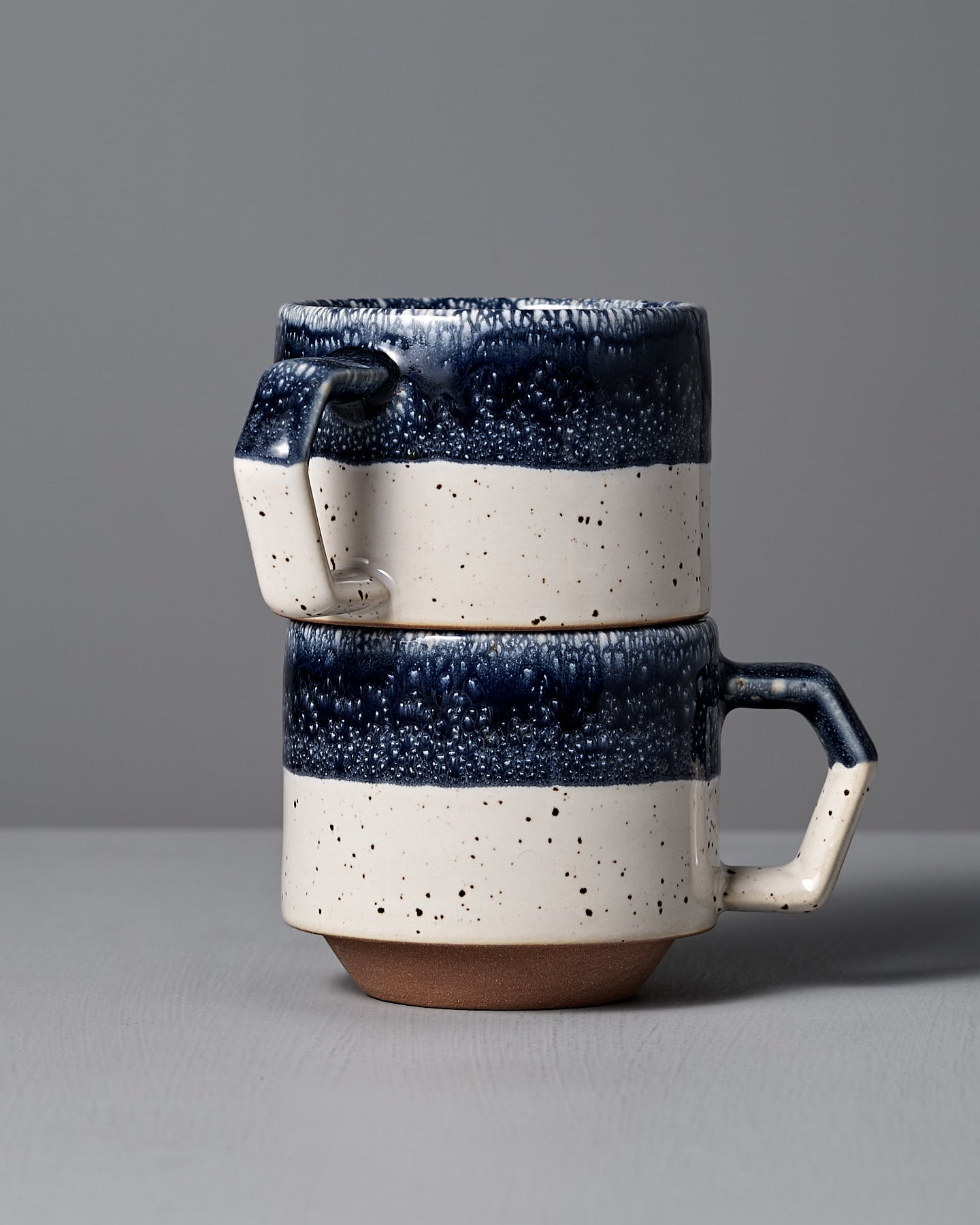 The Stacking Mug – White-Navy Drop by CHIPS Inc. features elegant Minoyaki porcelain with a textured dark blue top, speckled white bottom, and square handle. Its exquisite craftsmanship and stackable design make it a perfect collection addition, beautifully placed on any gray surface.